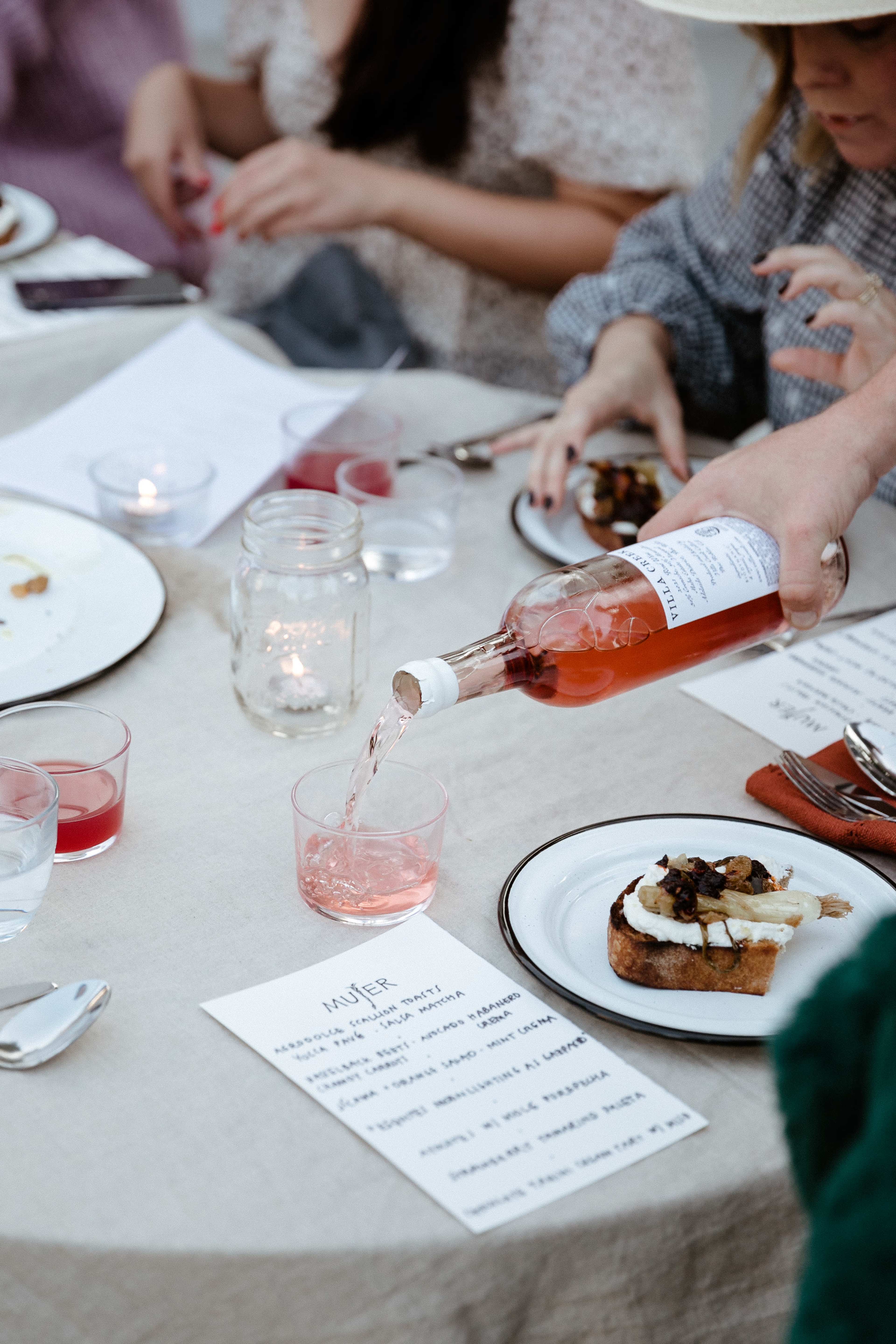 A person pours a pink drink from a bottle into a clear glass at a dining table set with food and notes.
