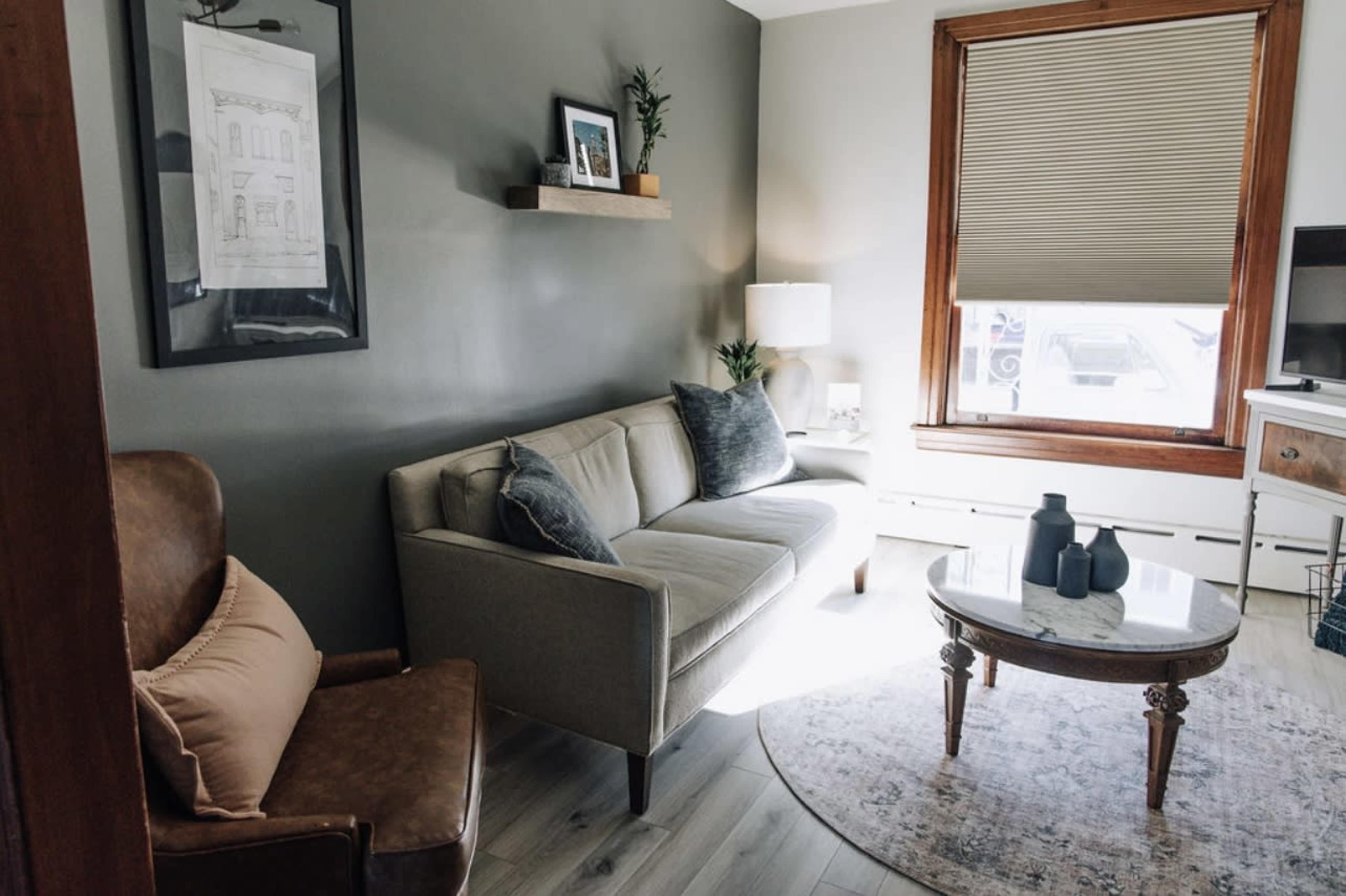 A living room with a gray wall, a light-colored sofa, a round coffee table, and a window with blinds.