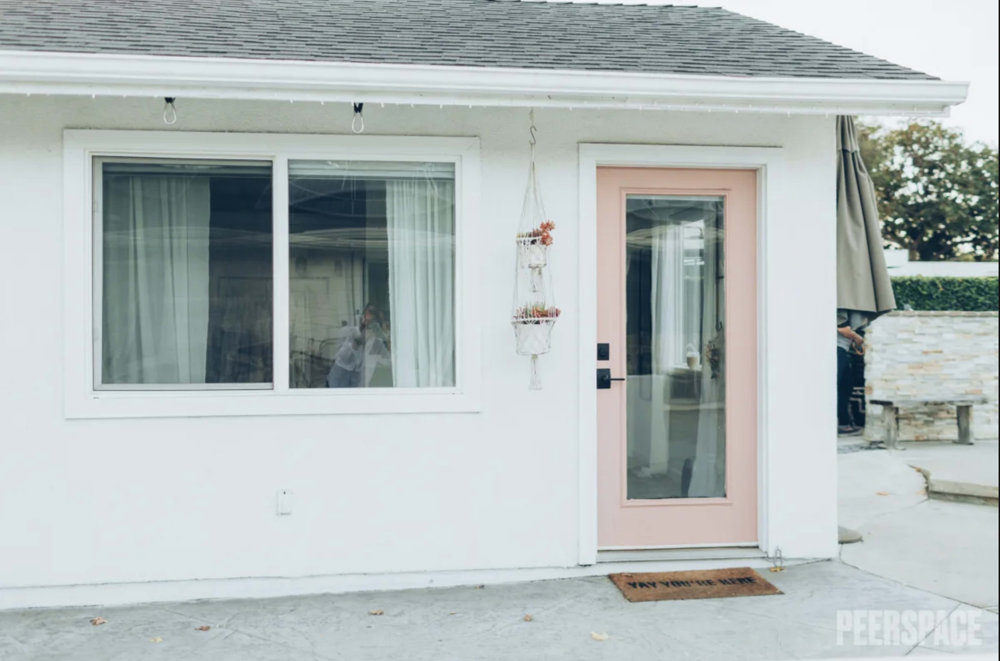 The image shows a white exterior of a small house with a pink door and a hanging planter by the entrance.