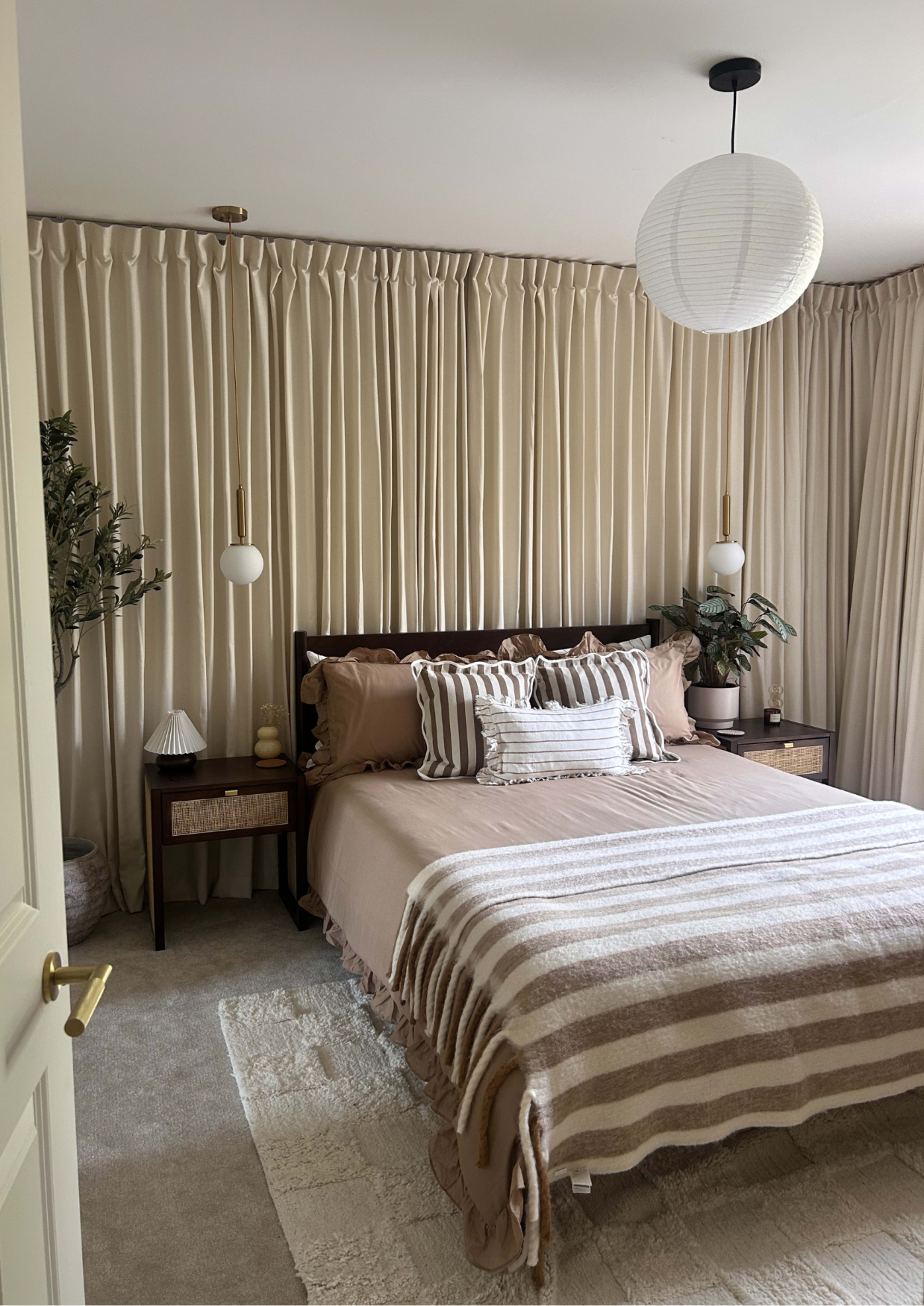 The bedroom features a beige color scheme with a bed neatly made with striped bedding, flanked by modern light fixtures and decorative plants.