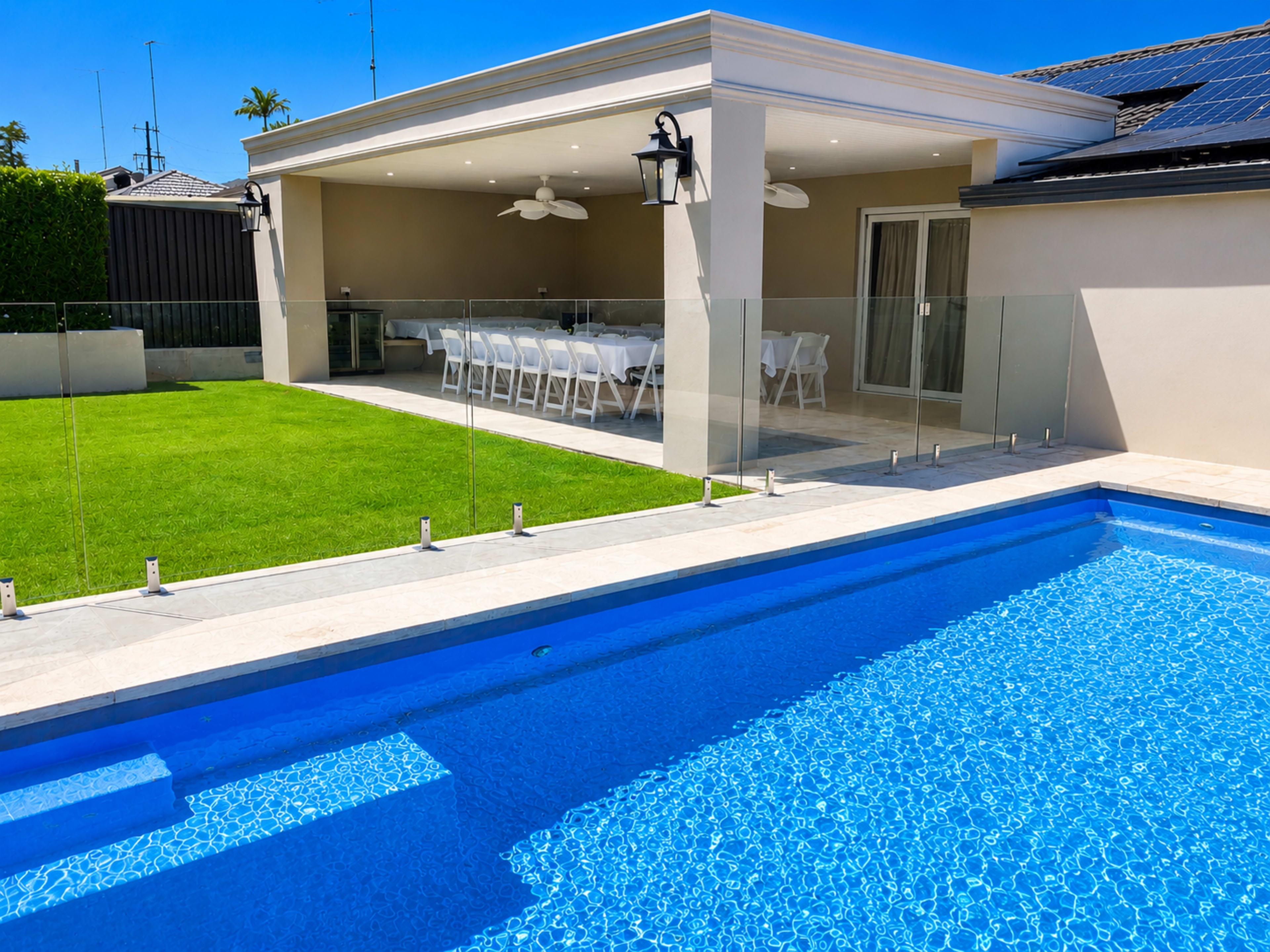The image shows a backyard with a swimming pool, a grassy area, and a covered outdoor dining space with a set of chairs and tables.