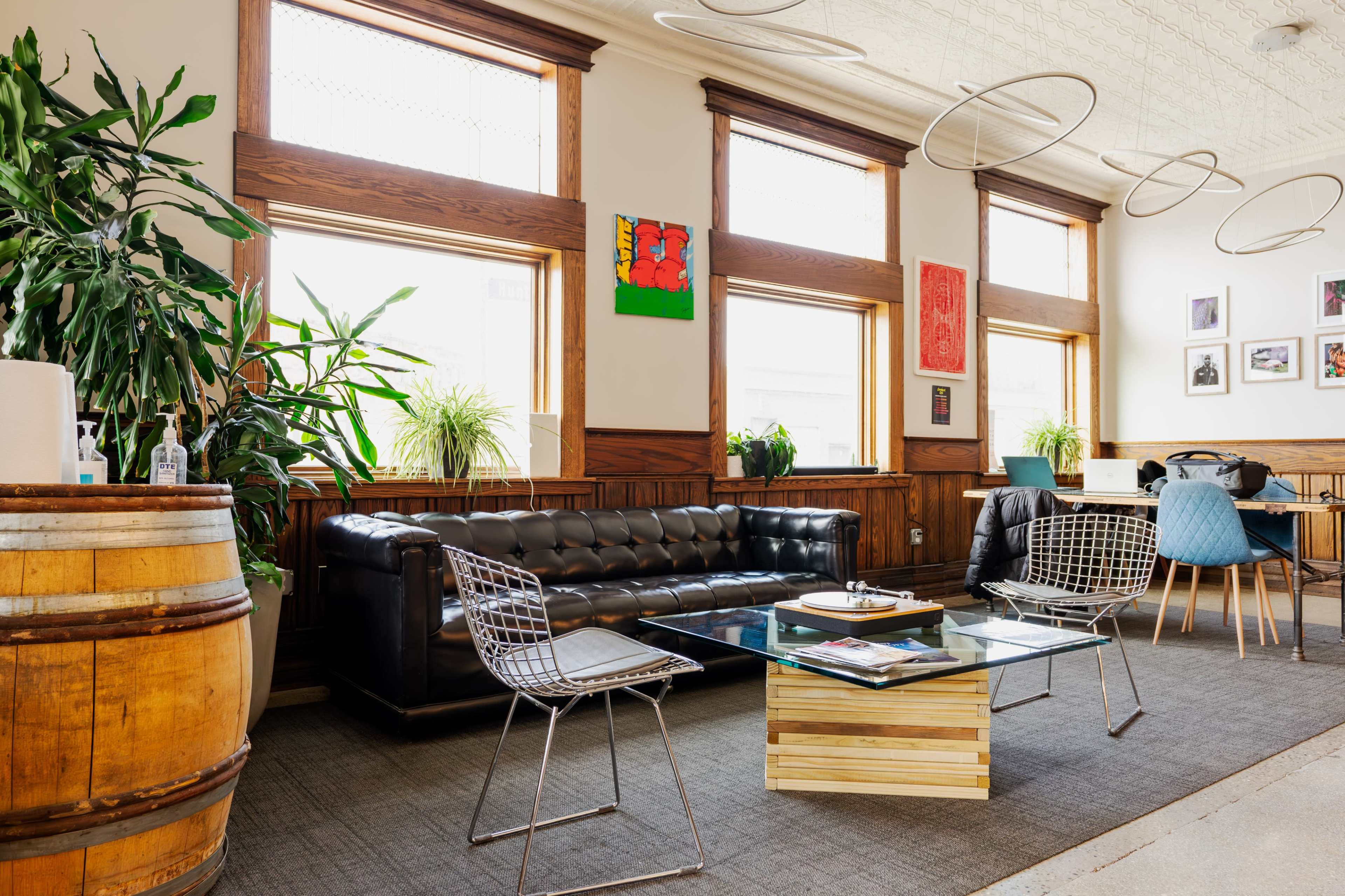 A modern, stylish lounge area featuring a black leather sofa, a glass coffee table, wooden barrel decor, and several plants, with artwork displayed on the walls.