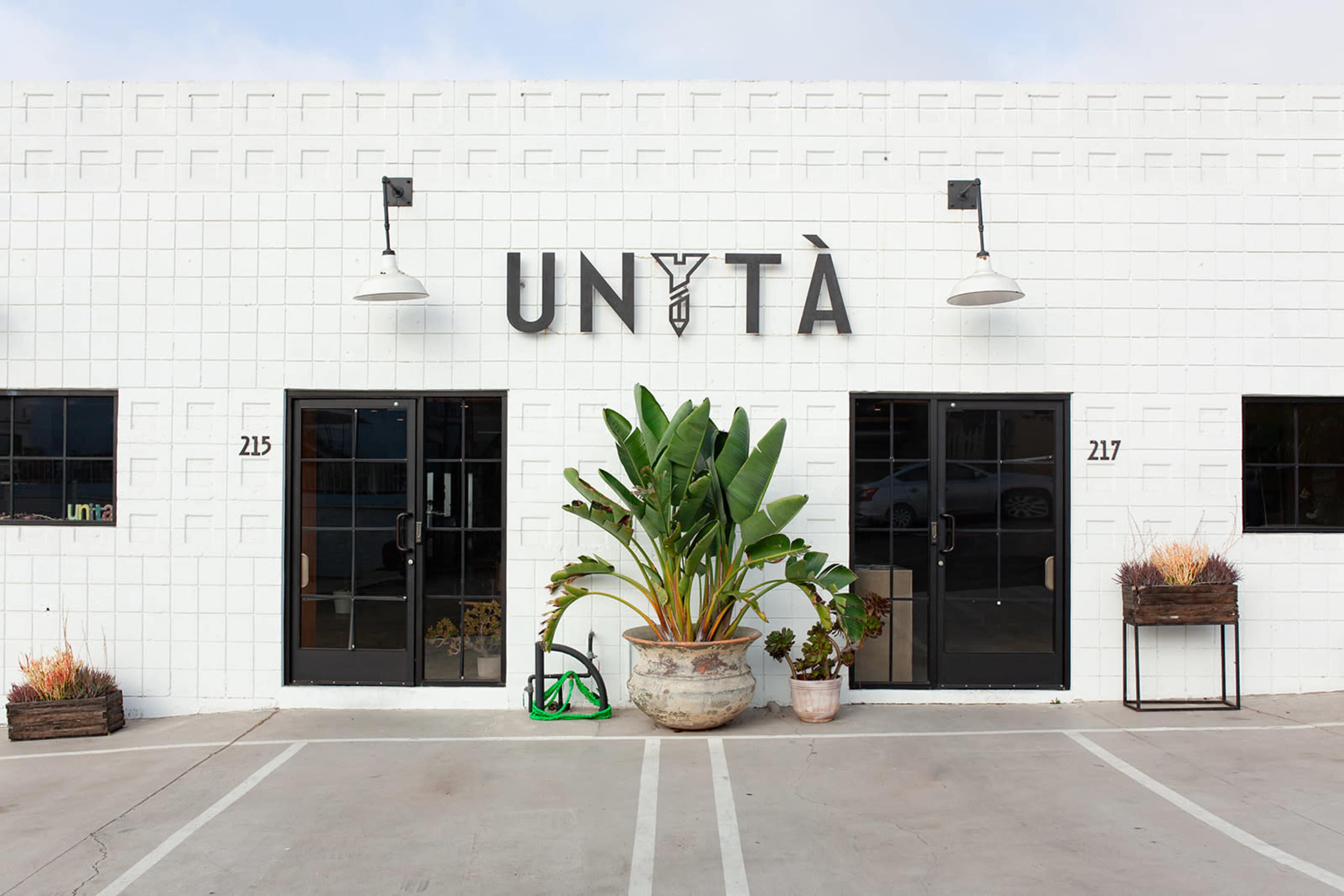 The image shows the exterior of a modern building with a white facade, featuring a large sign that reads "UNITY" and decorative plants near the entrance.