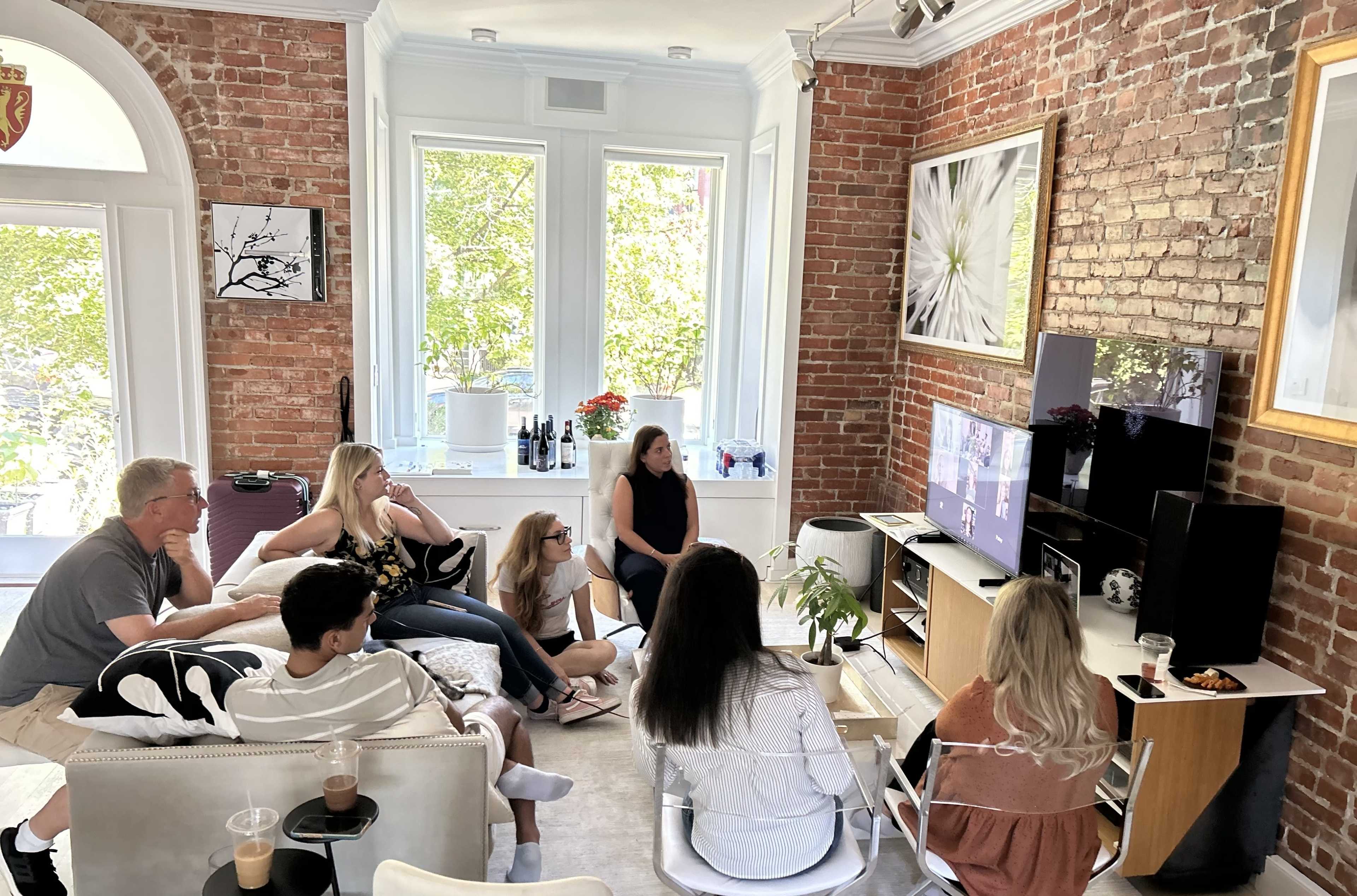 A group of people is sitting in a living room with exposed brick walls, watching a television screen while engaging in conversation.