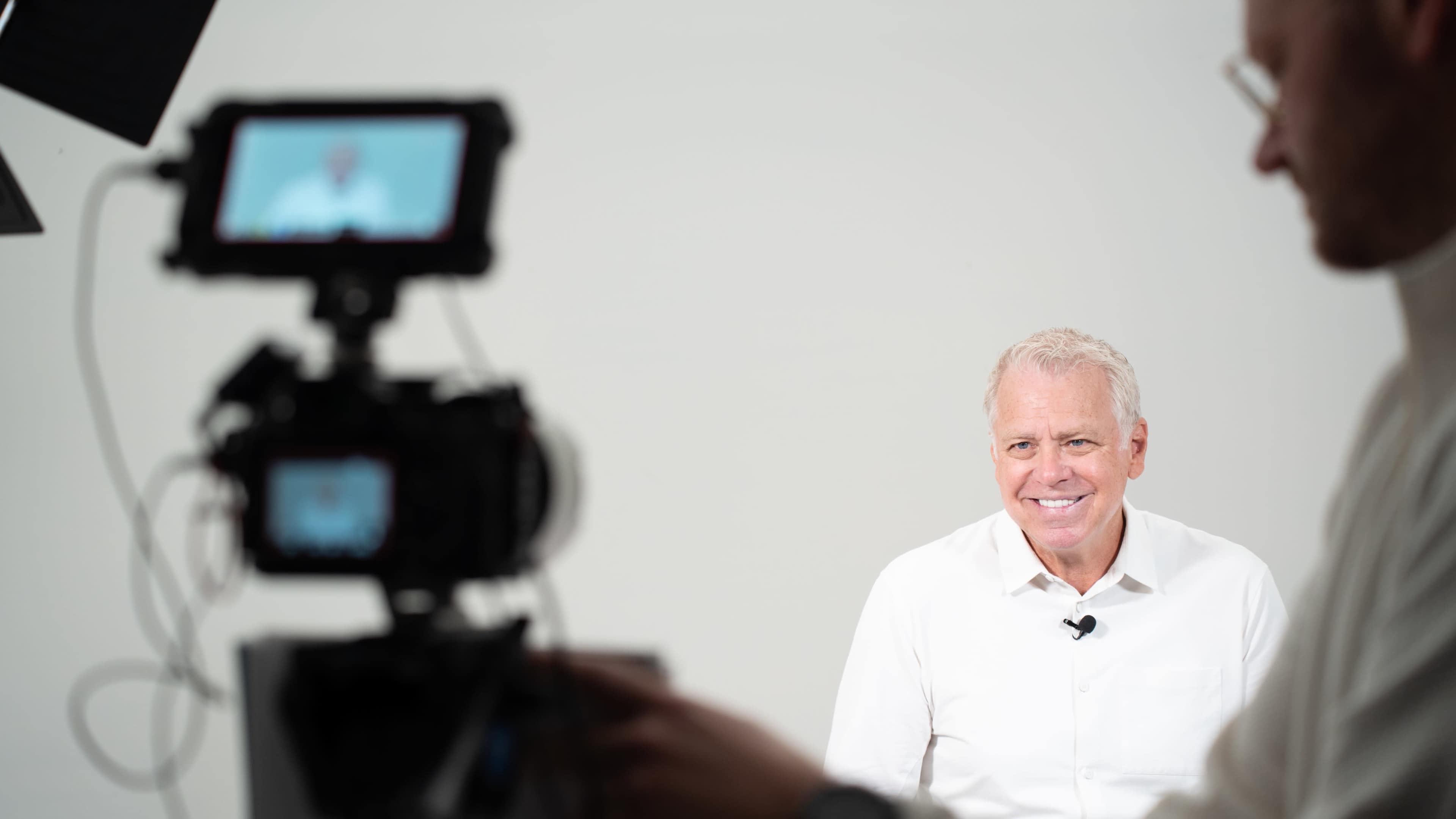A man in a white shirt is smiling while sitting in front of a camera on a tripod.