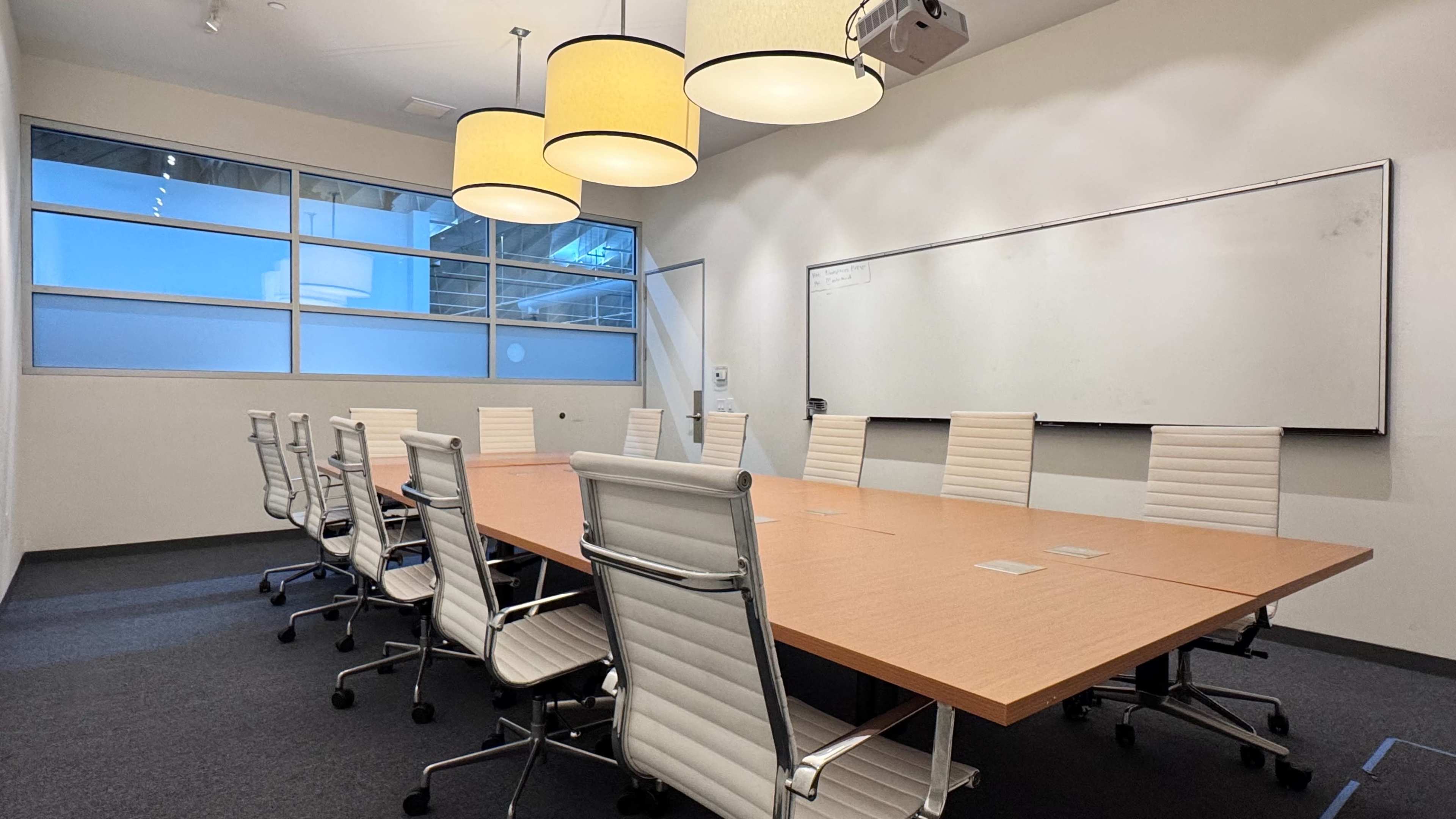 The image shows a modern conference room with a large wooden table, several white office chairs, and overhead pendant lights.