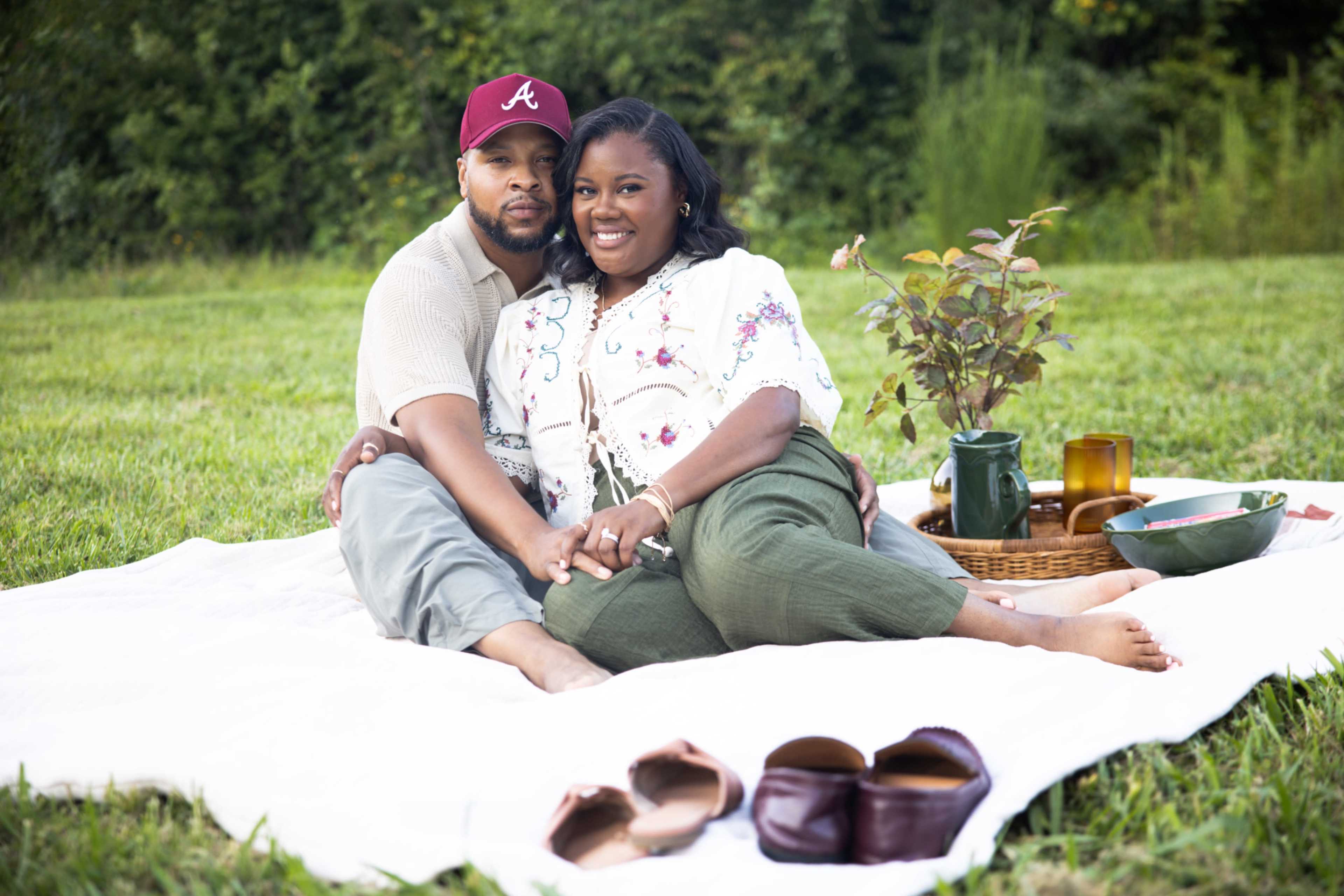 A couple sits on a white blanket in a grassy area, surrounded by various picnic items and greenery.
