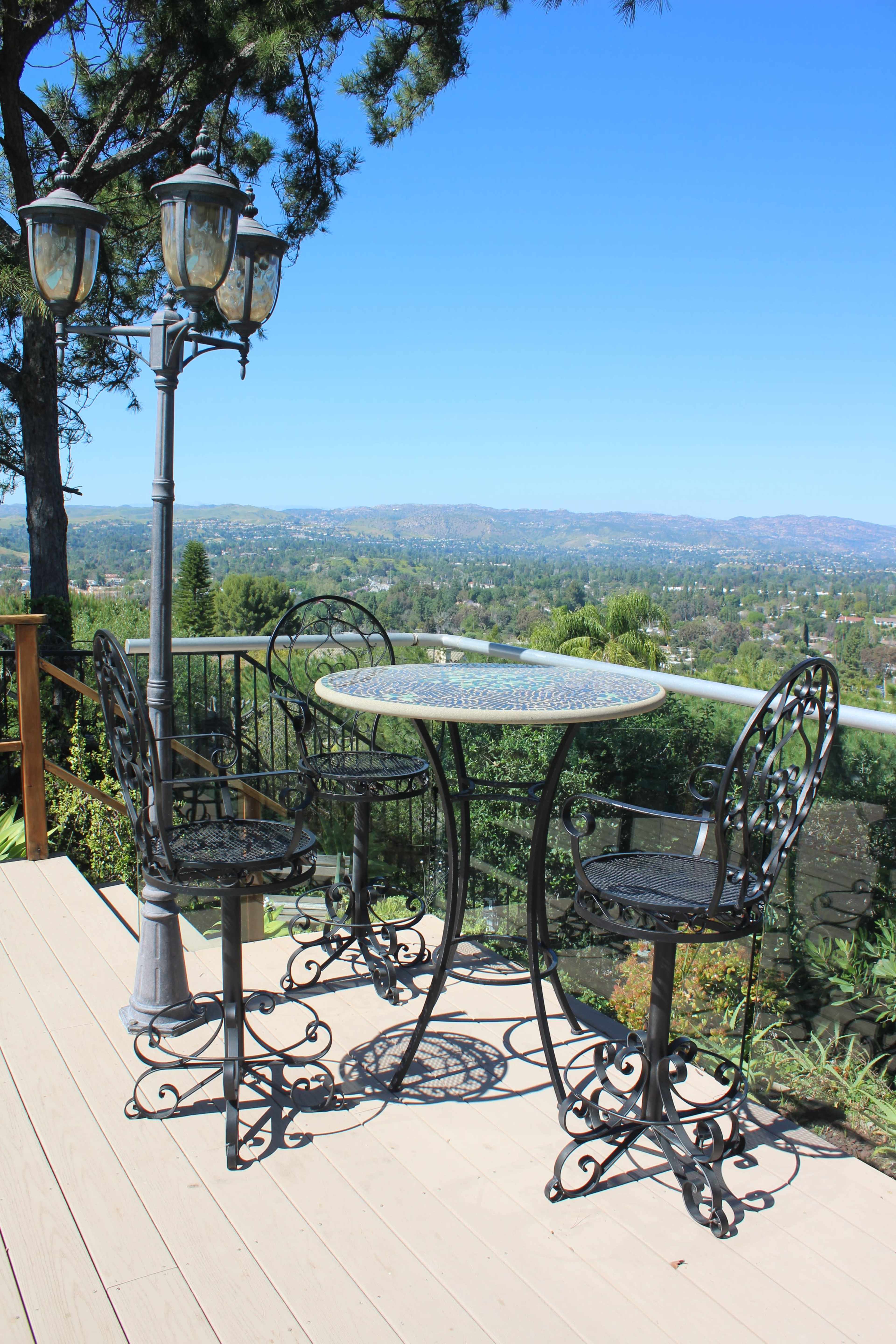 A wrought iron table and two chairs are positioned on a wooden deck overlooking a lush valley and blue sky.