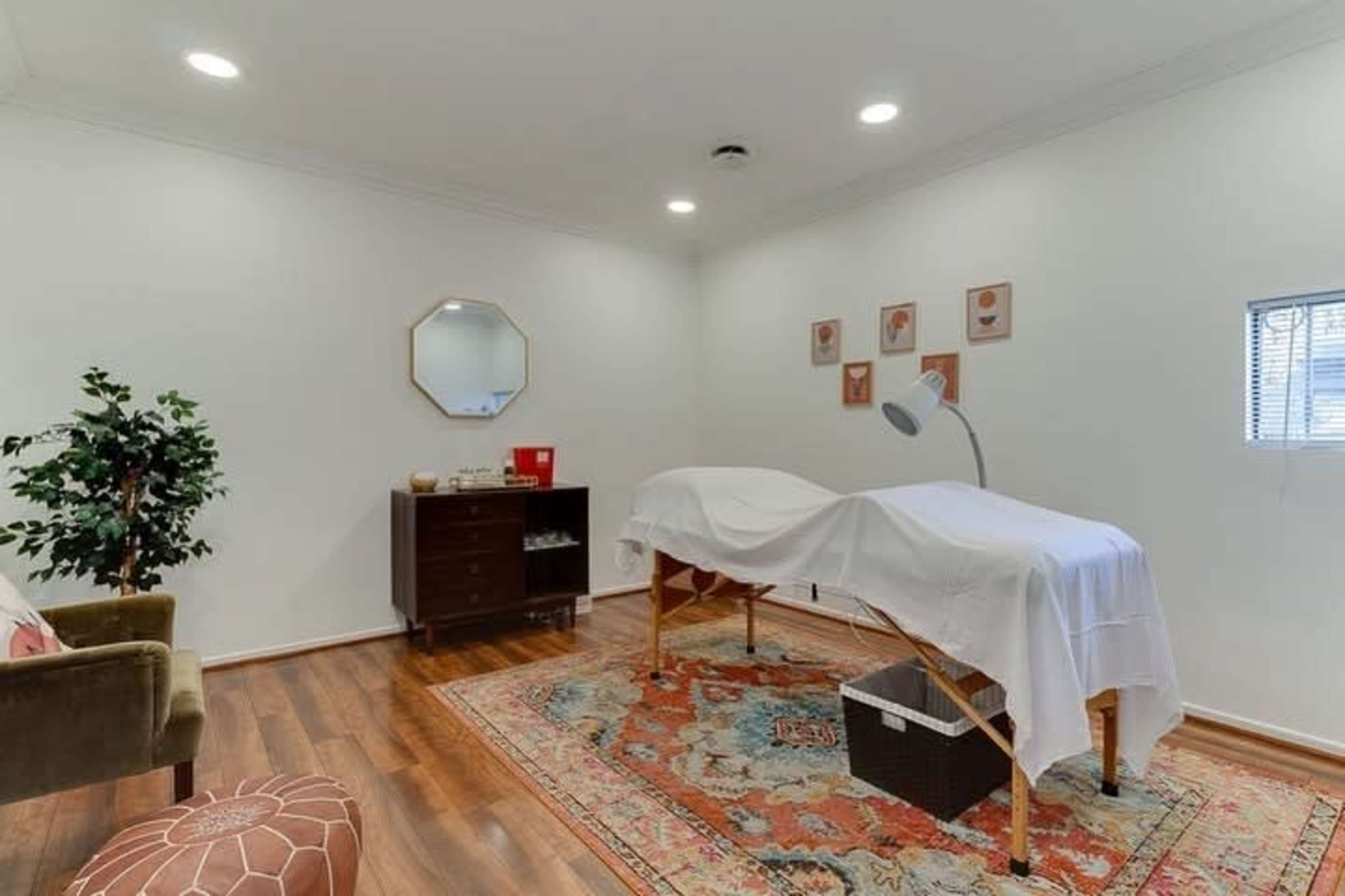 The image shows a treatment room with a massage table draped in white linens, a wooden storage unit, a mirror, and wall art arranged above the table.