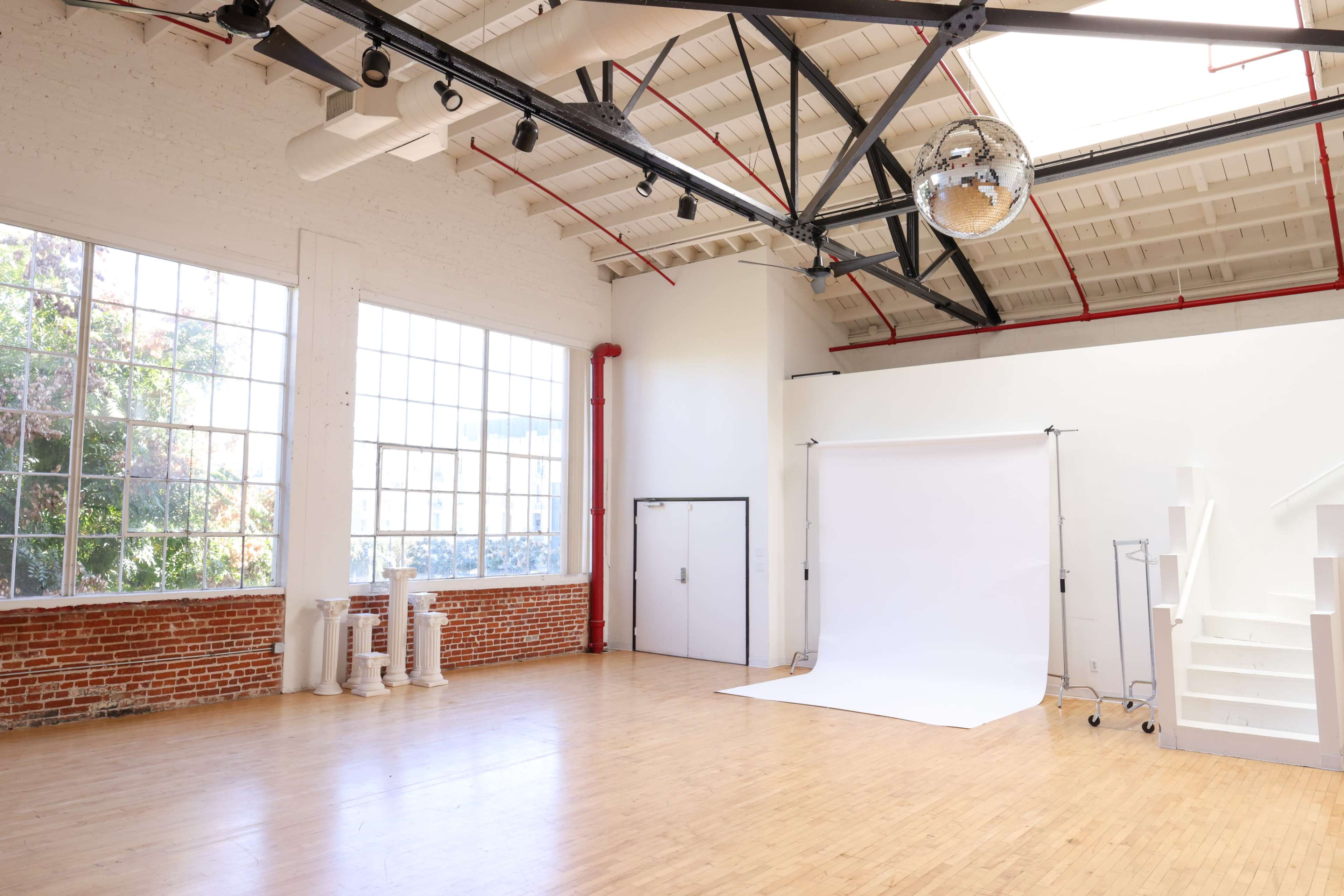 A spacious, well-lit studio features large windows, polished wooden floors, a white backdrop on a stand, and a disco ball hanging from the ceiling.