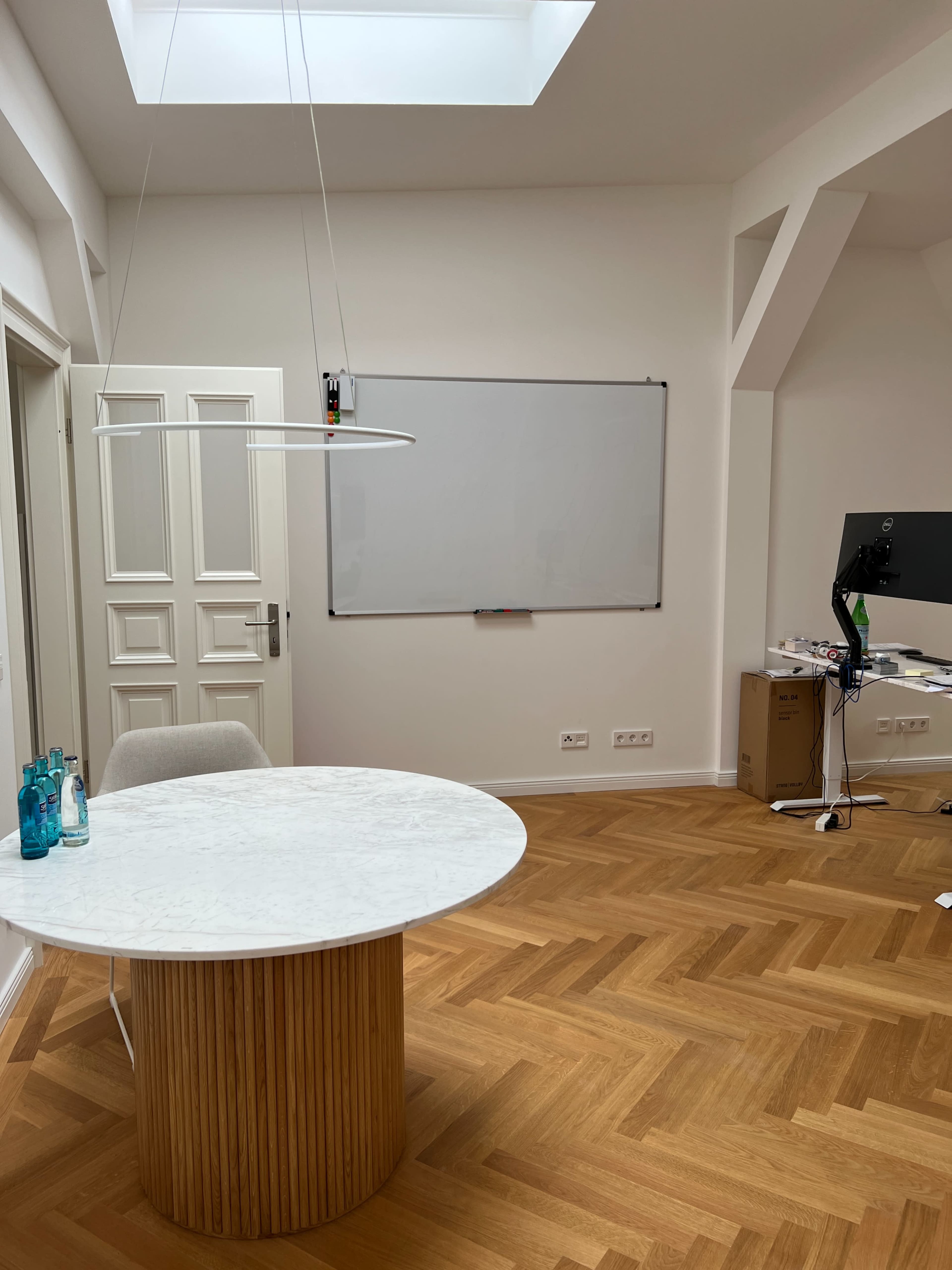 A minimalist office with a round white table, a whiteboard, a computer setup, and a door leading to another room.