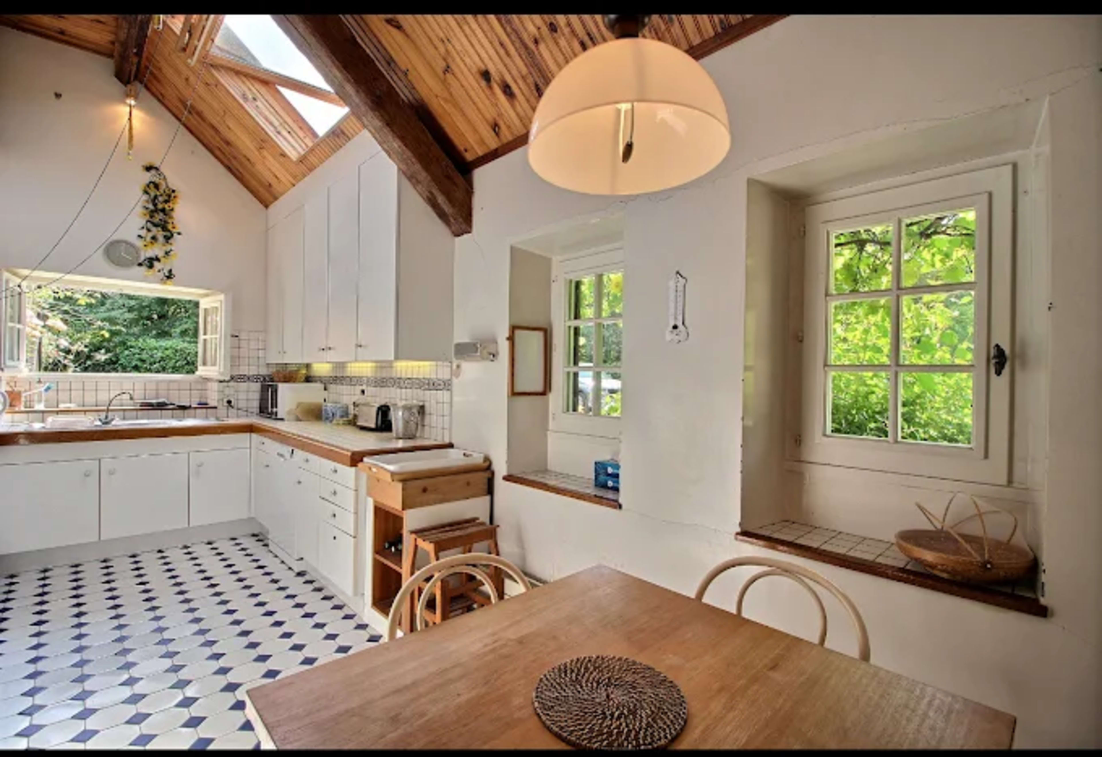 The image shows a bright kitchen with a wooden ceiling, white cabinets, a wooden table, and a window with leafy greenery outside.