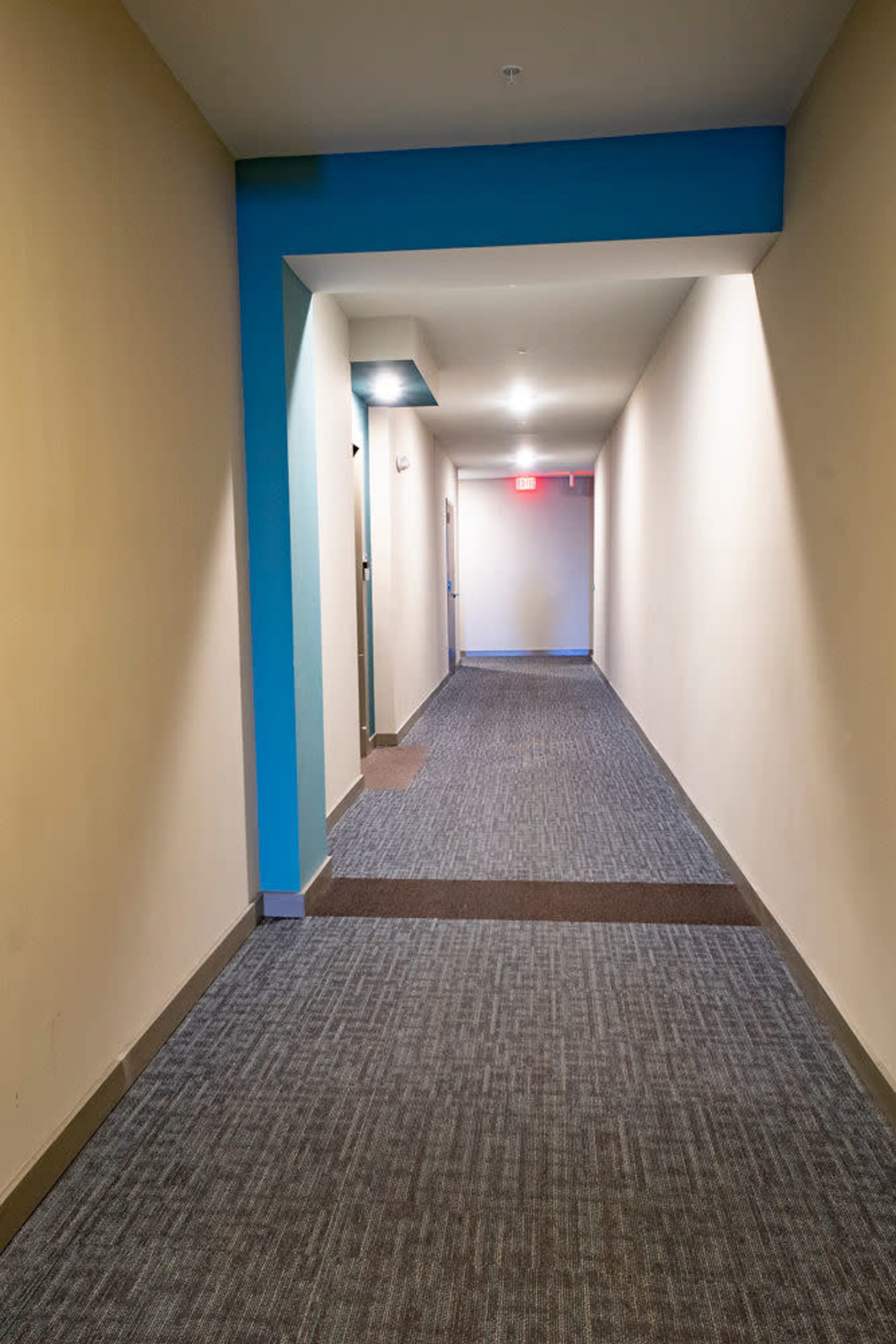 A long, narrow hallway with gray carpet, beige walls, and a blue accent on the ceiling.