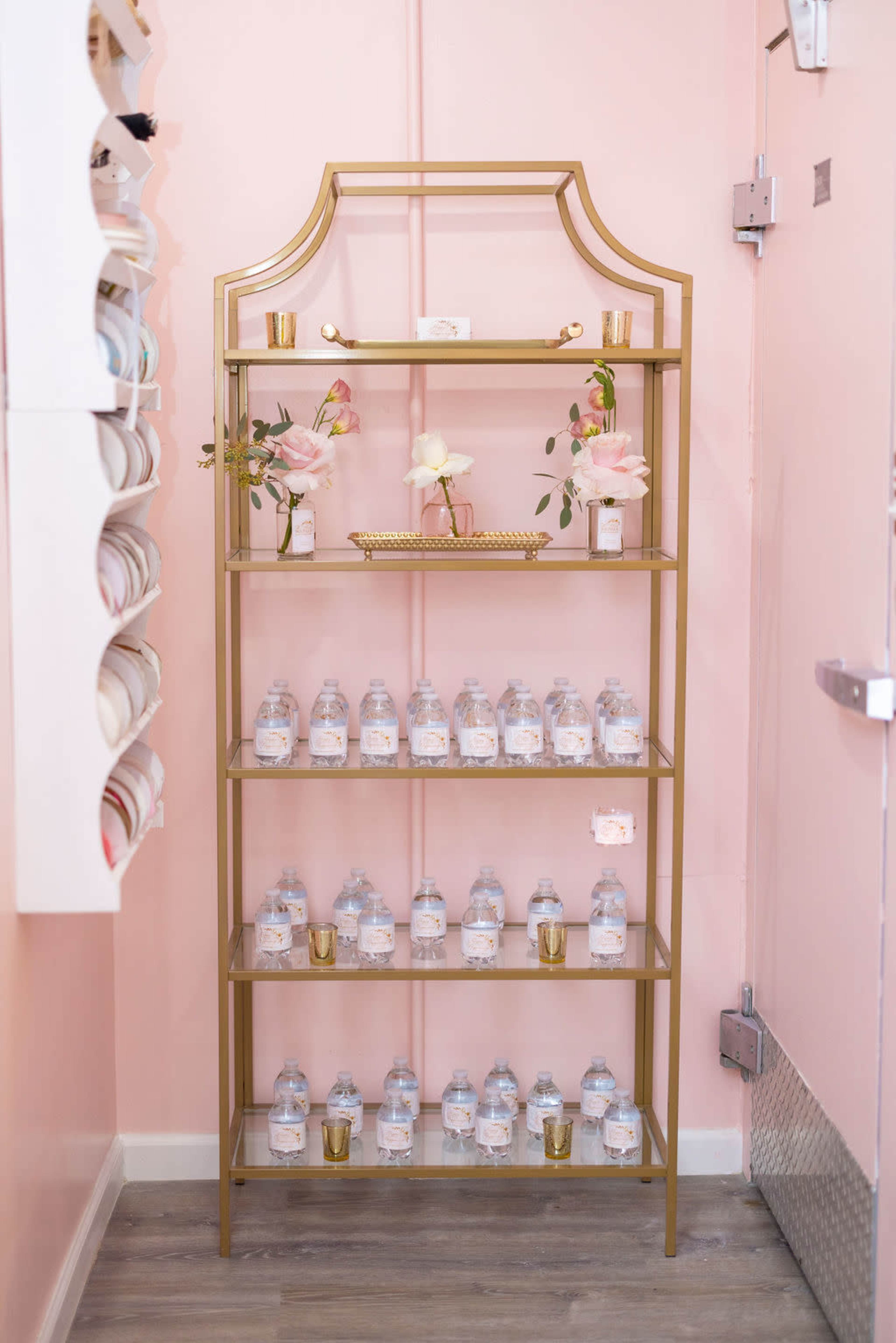 A gold shelving unit displays water bottles and decorative items against a pink wall.