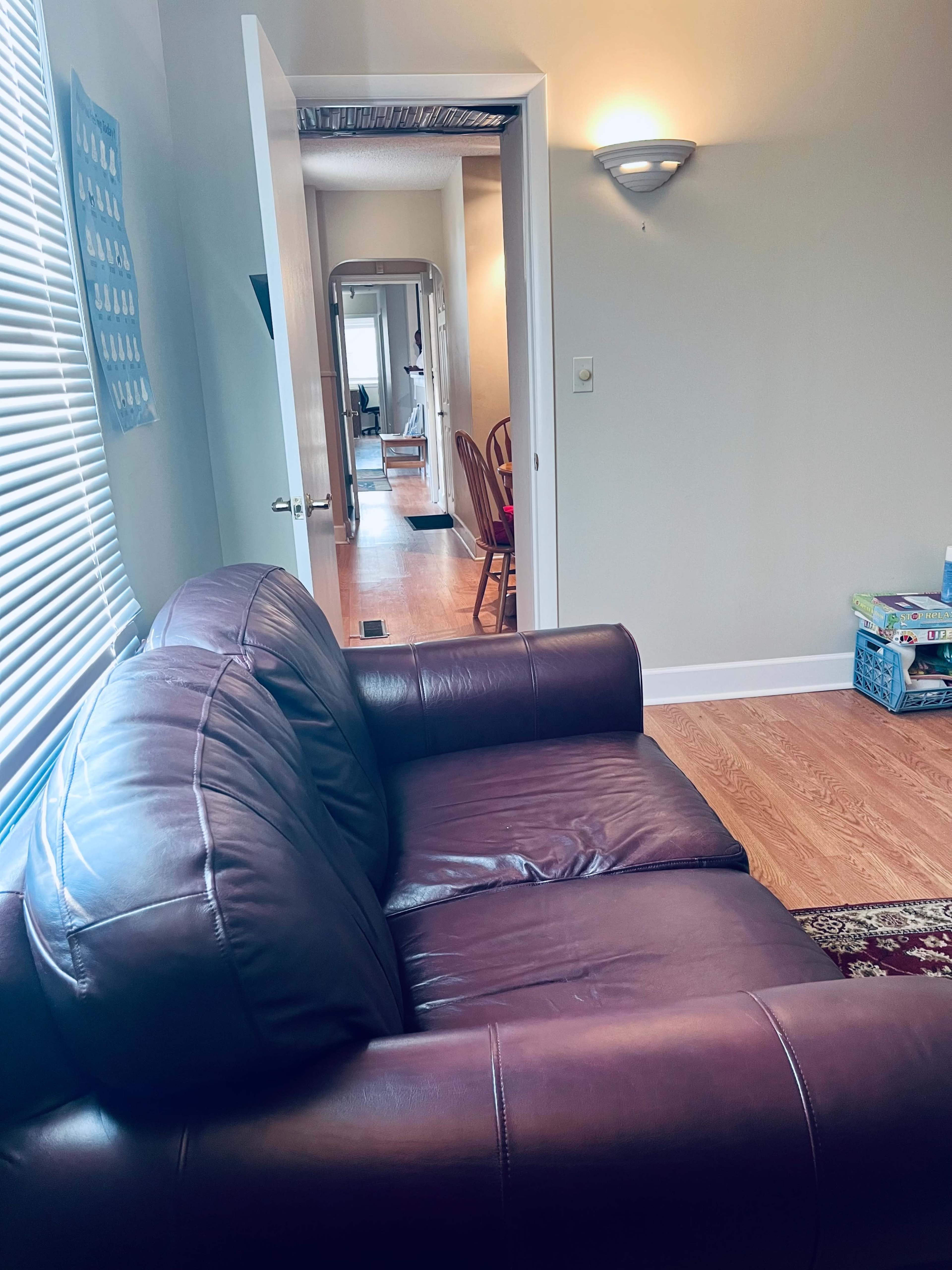 A brown leather couch is positioned in a well-lit living room, facing an open door that leads into a hallway.