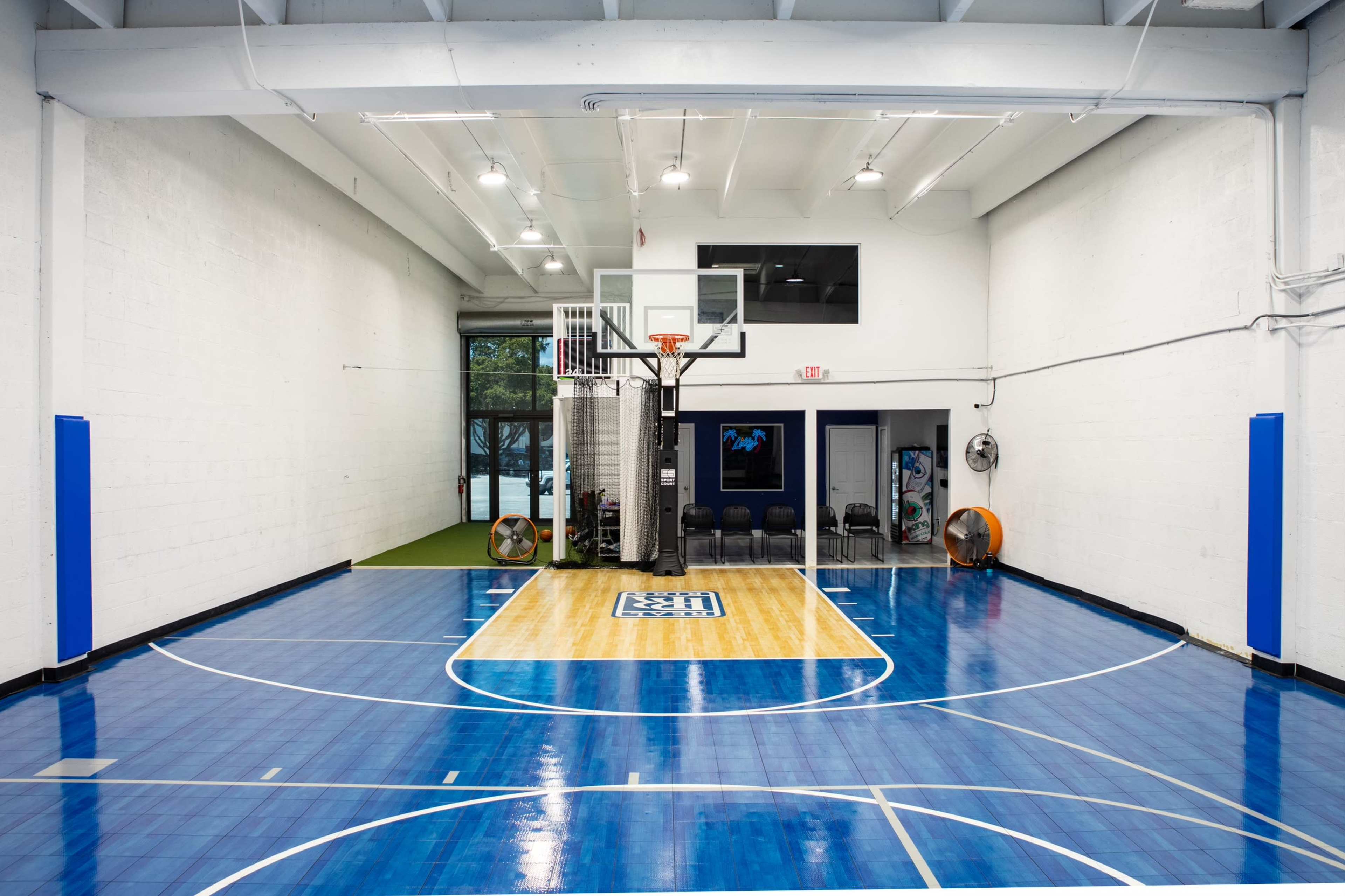 A basketball court with a polished wooden center and blue flooring, flanked by white walls and seating along one side.