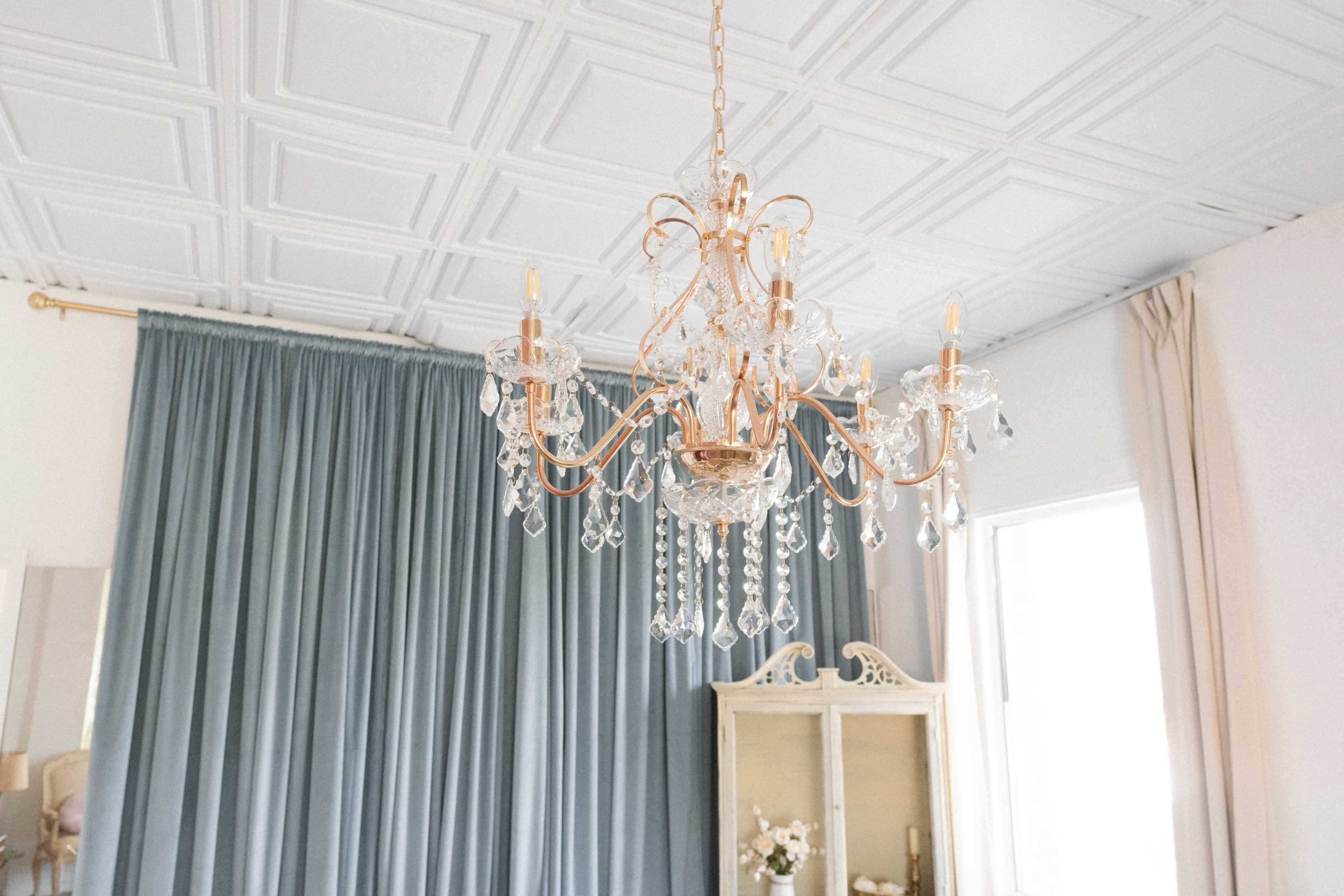 A crystal chandelier hangs from the ceiling above a room with blue curtains and a light-colored cabinet.