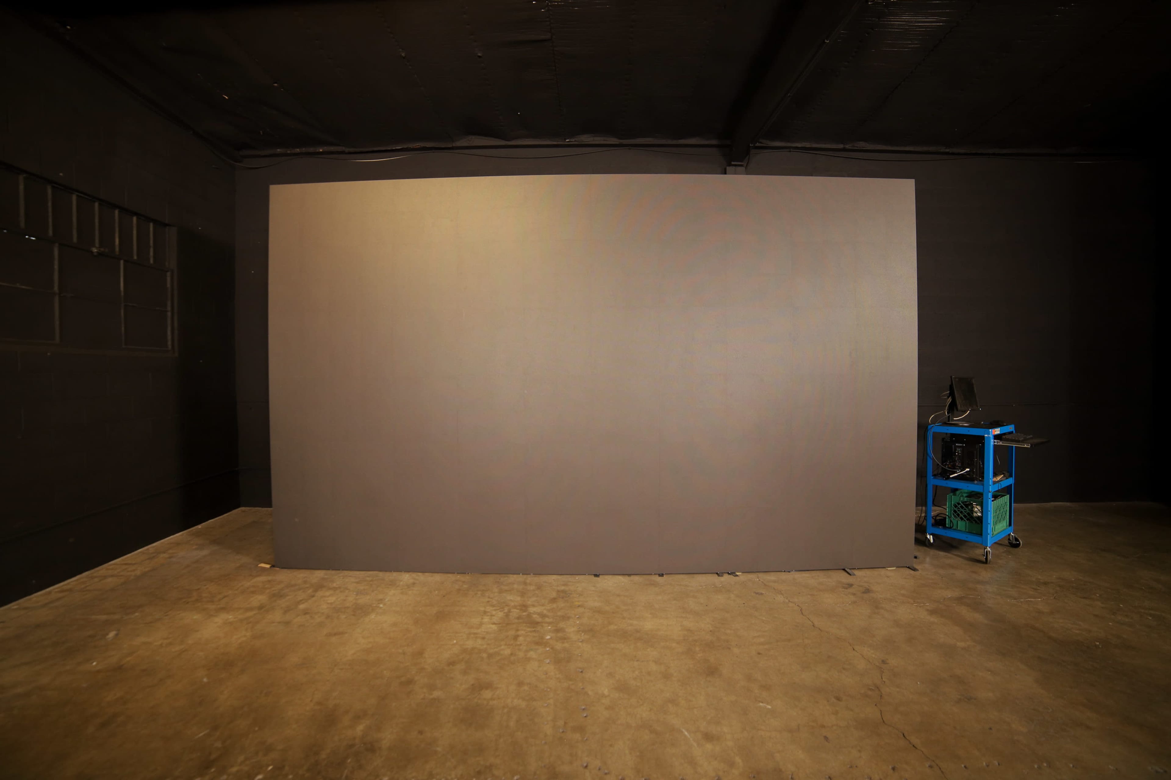 A large, blank gray wall in a dimly lit room with a concrete floor and a blue cart on the right side.