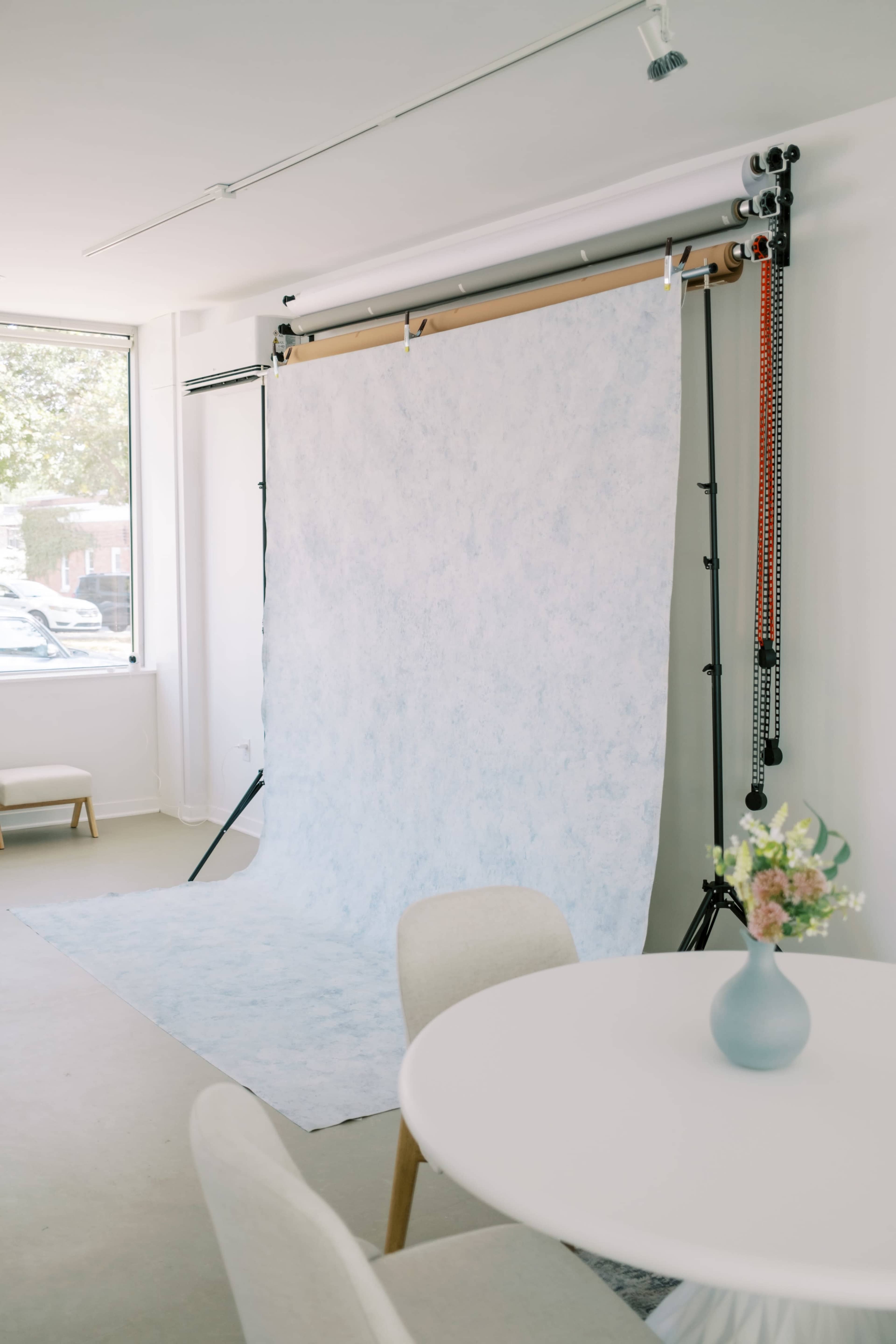 The image shows a photography studio featuring a light blue backdrop on a support stand, a round table with a flower vase, and a chair in a bright, minimalist space.