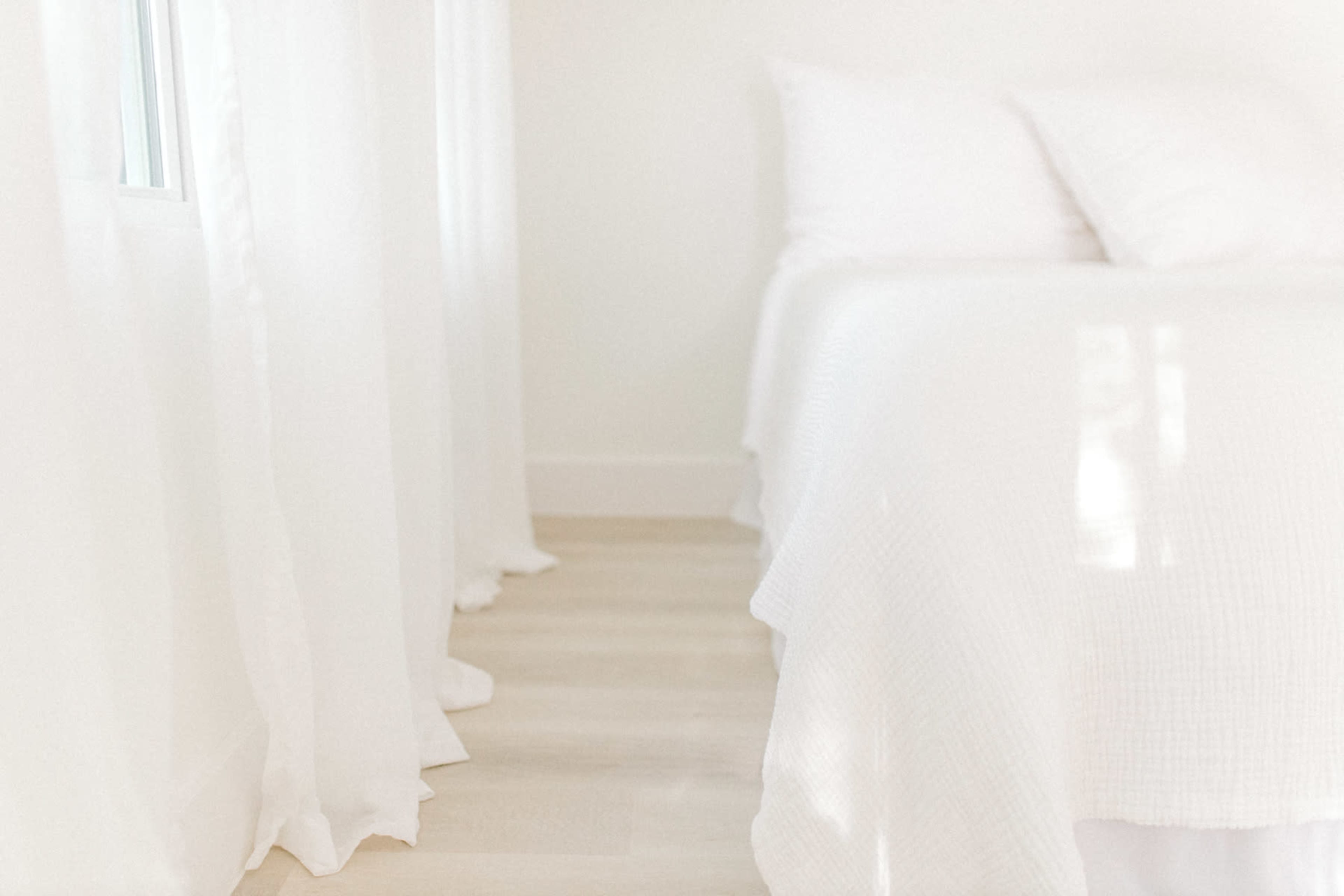 The image shows a light, minimalistic bedroom with a bed and sheer white curtains.