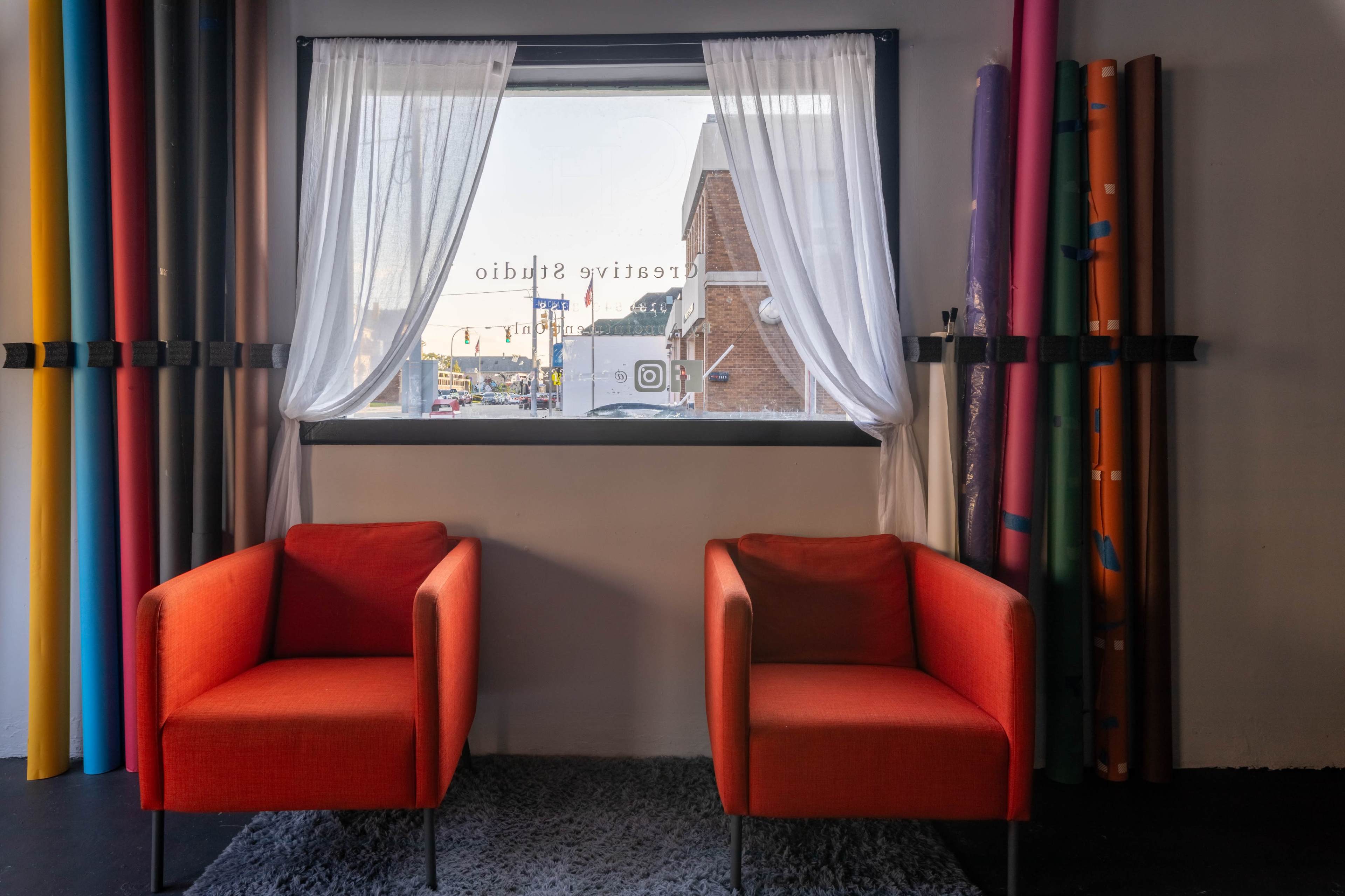 Two orange chairs are positioned in front of a large window, adorned with sheer white curtains, overlooking a street scene with colorful rolls of backdrop material displayed nearby.