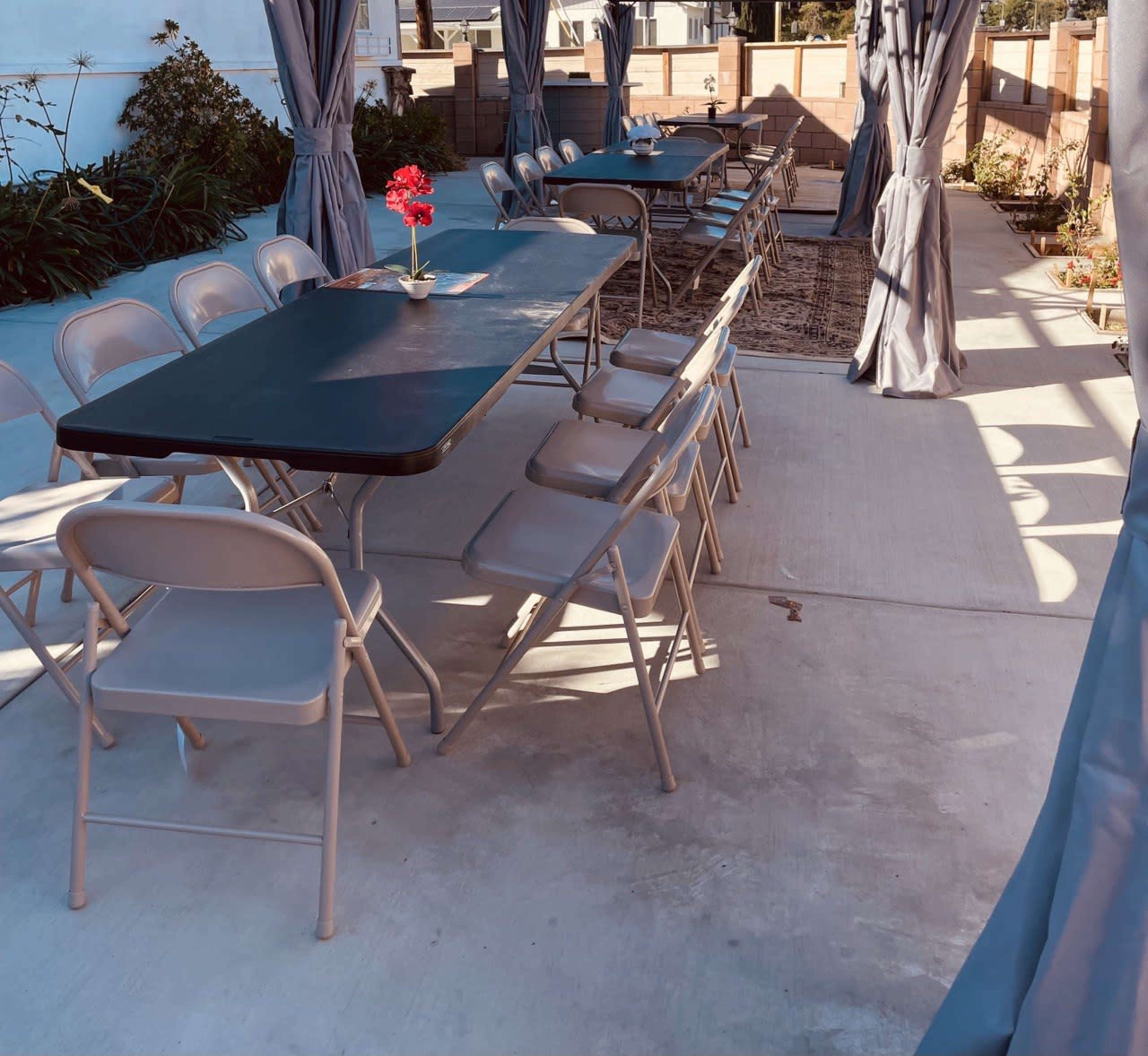 A covered outdoor seating area features several metal tables and folding chairs arranged on a concrete surface, with a single potted flower on one table.