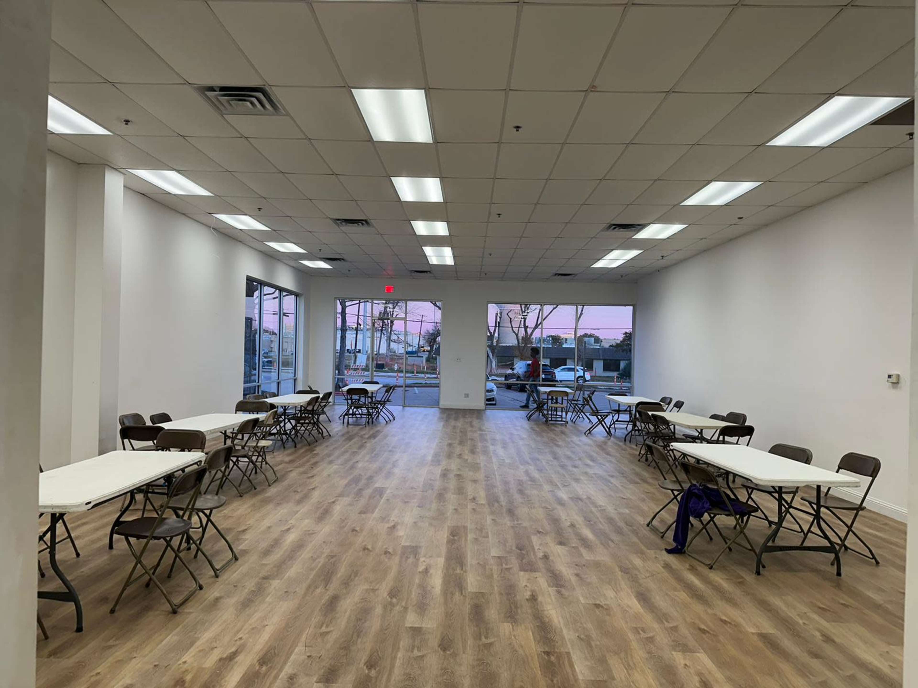 A spacious, well-lit room with multiple long tables set up with chairs and large windows revealing a sunset view outside.