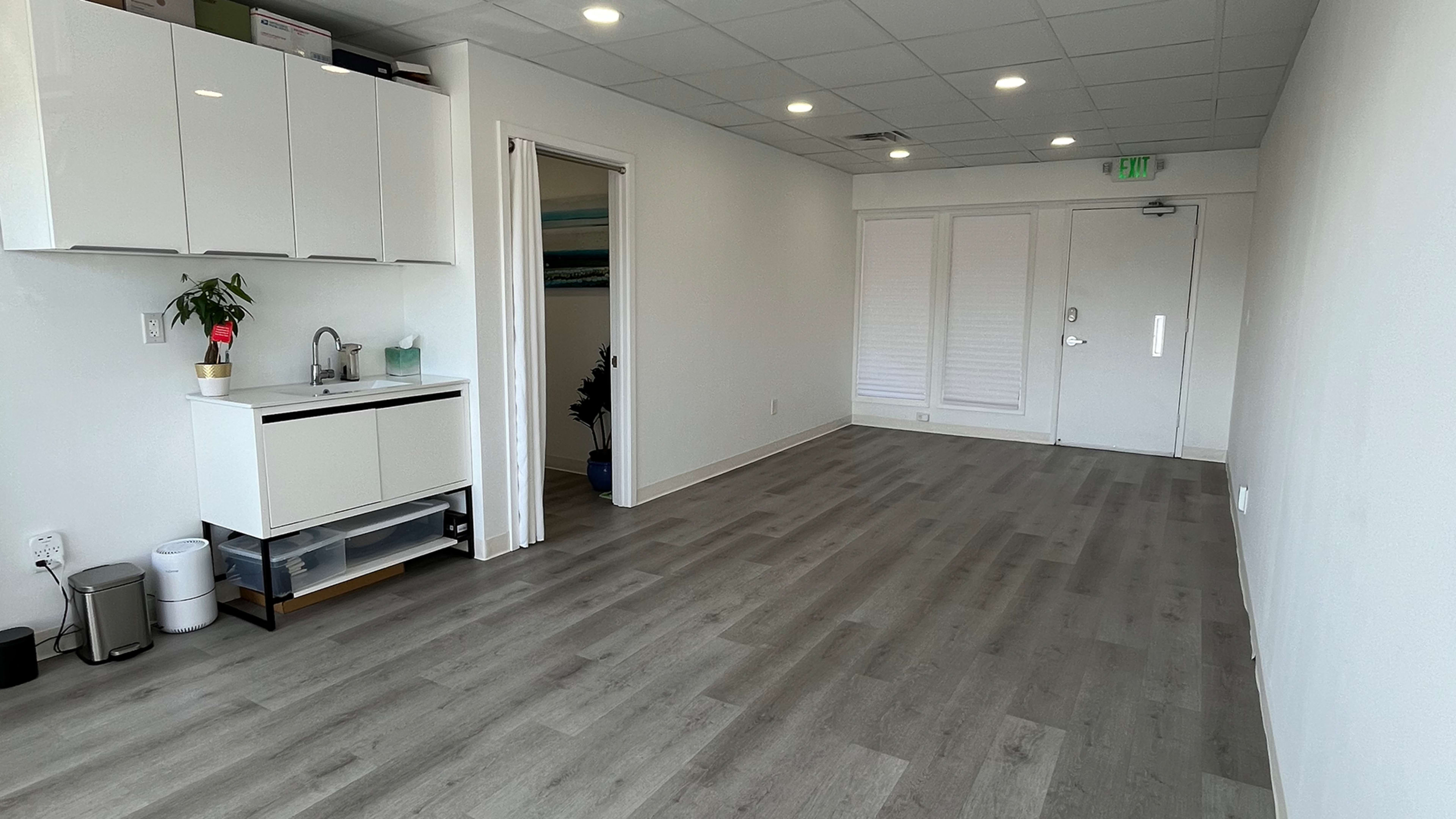 The image shows a well-lit, empty room with a small kitchenette on one side and sliding glass doors on the opposite side.