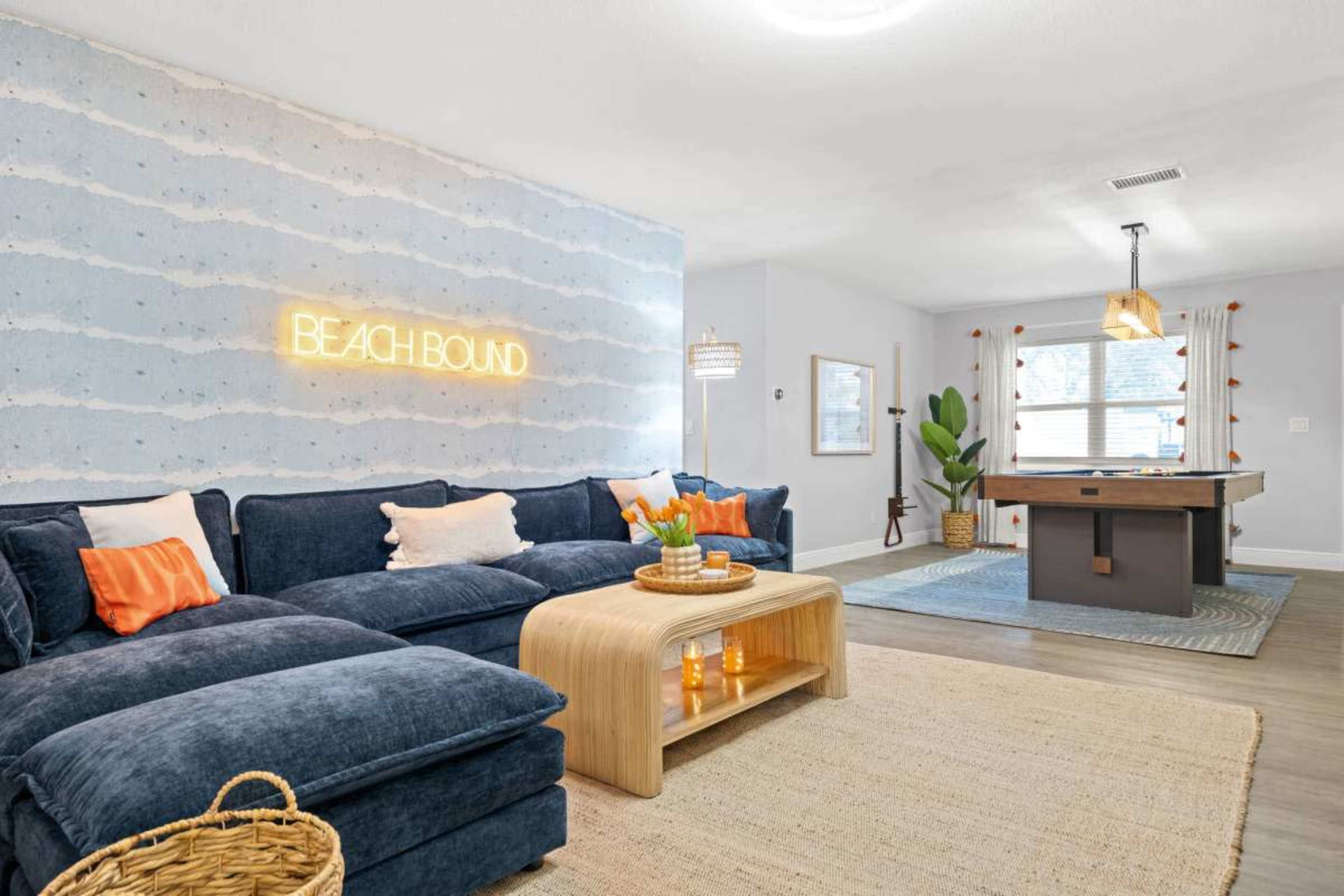 The image depicts a modern living room featuring a blue sectional sofa with orange accent pillows, a wooden coffee table, and a neon sign that says "BEACH BOUND" on the wall.