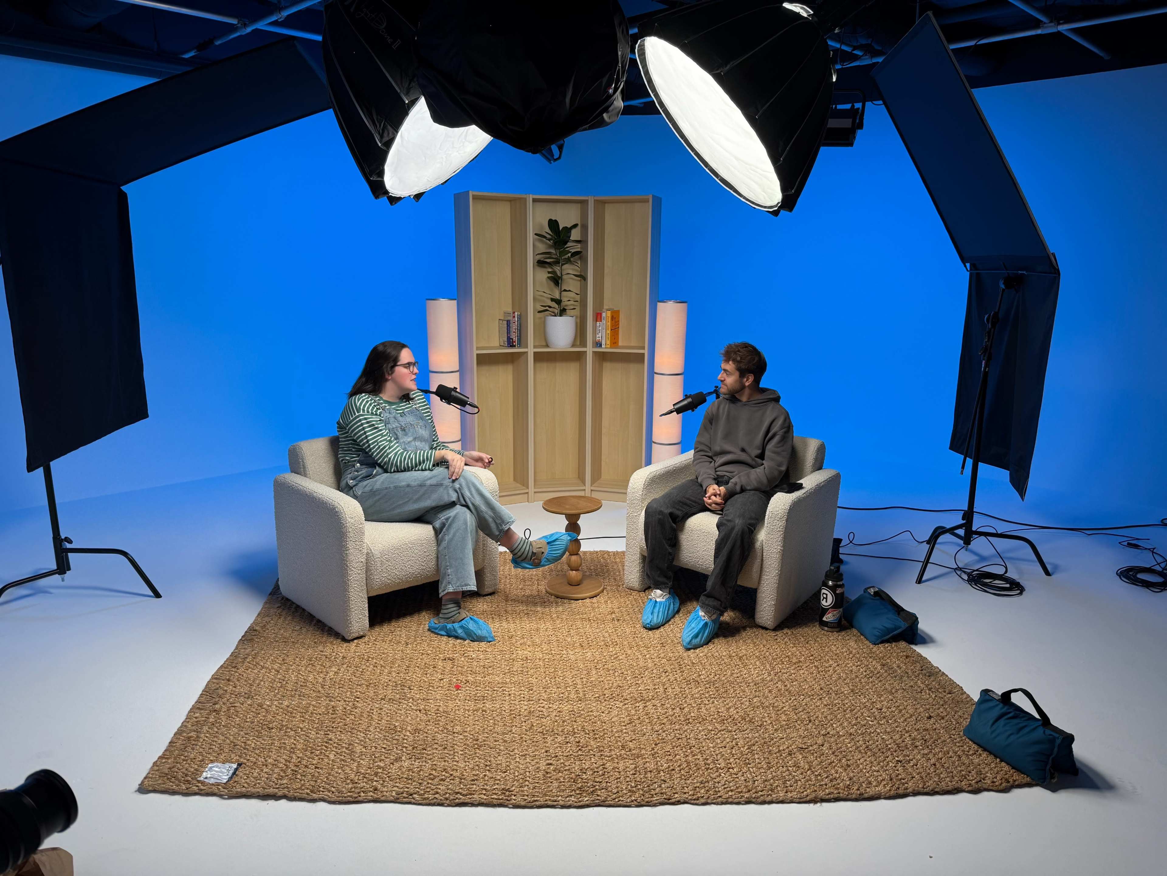 A podcast setting features two people seated in armchairs facing each other, with microphones, large light sources overhead, and a minimalist background.