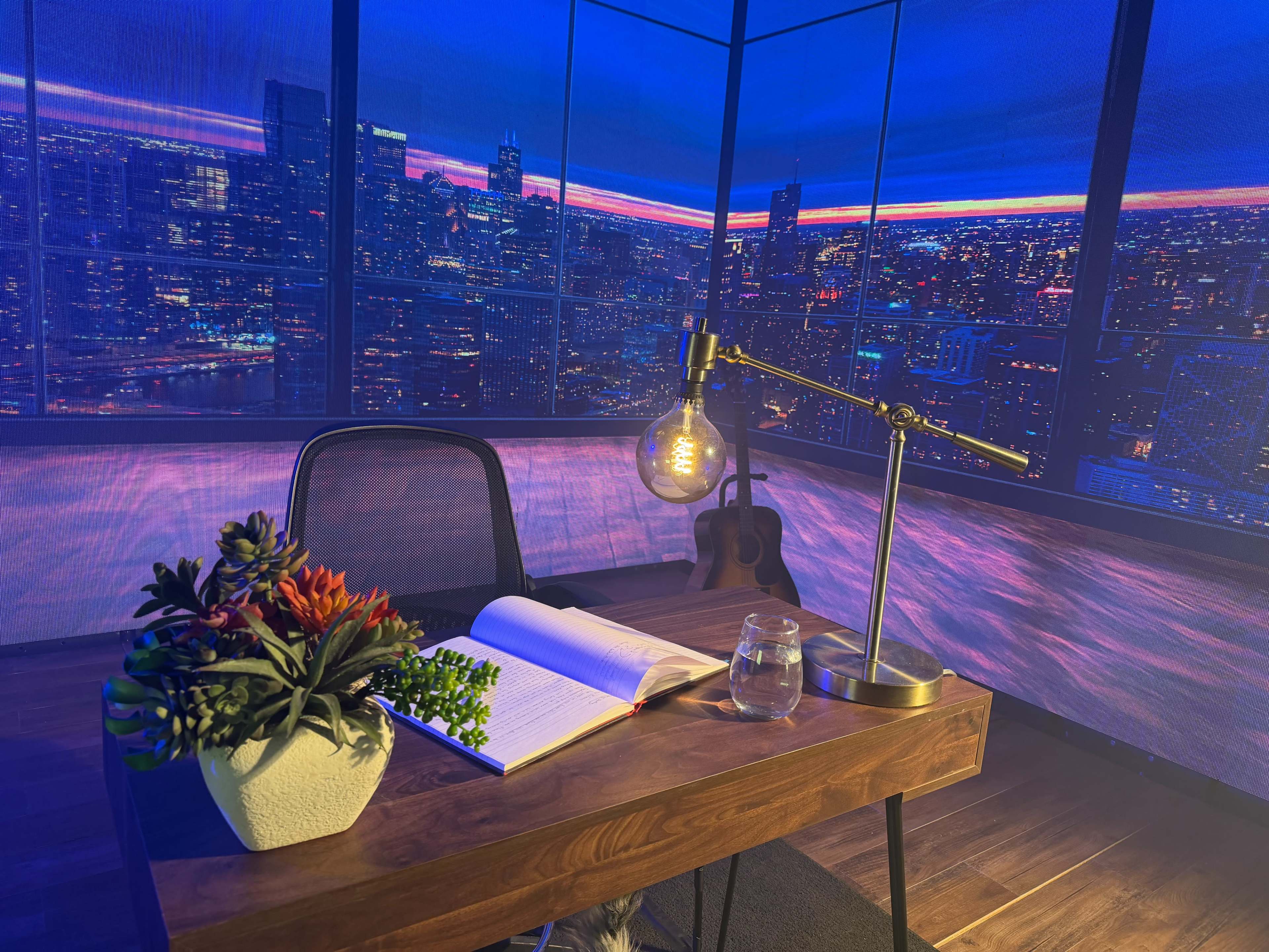 A desk with a lamp, an open book, a glass jar, and a potted plant is set against a backdrop of a city skyline at night.