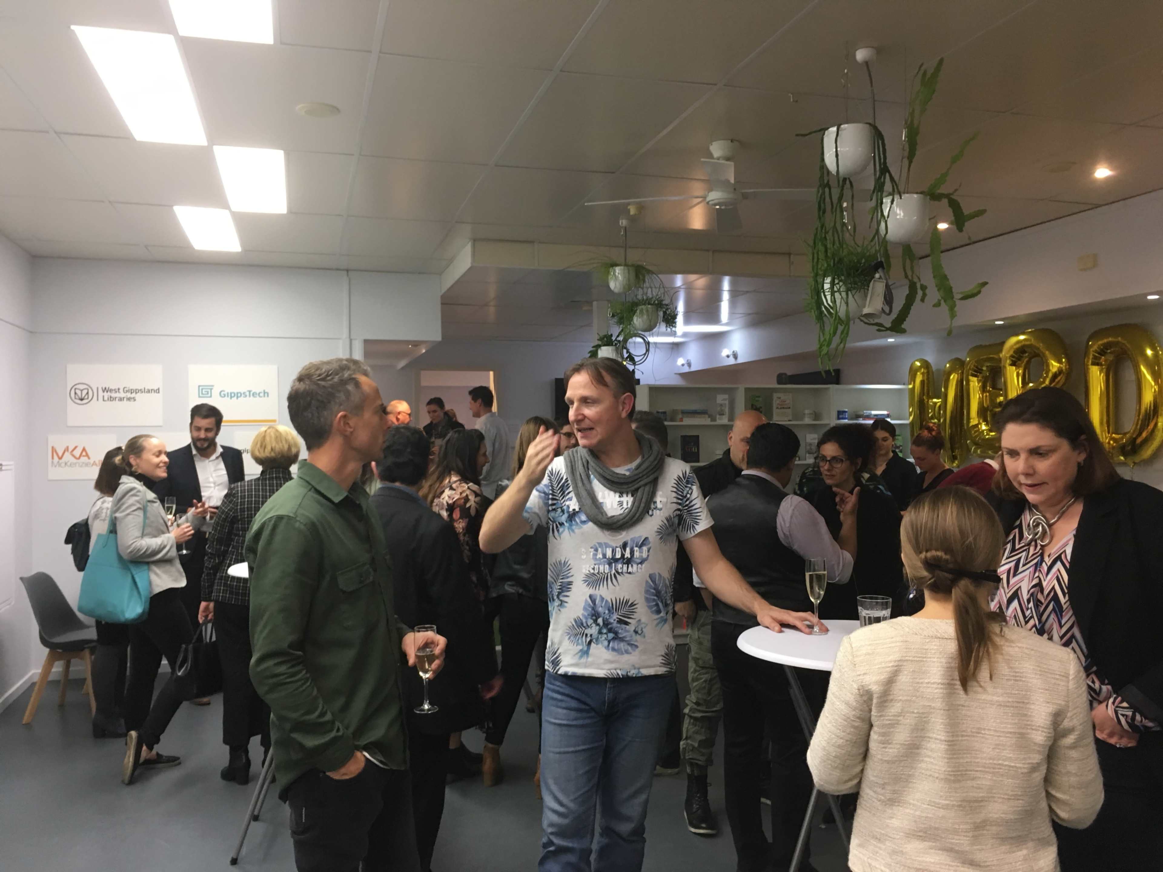 A group of people socialize at a lively event, standing around high tables in a well-lit room adorned with plants.