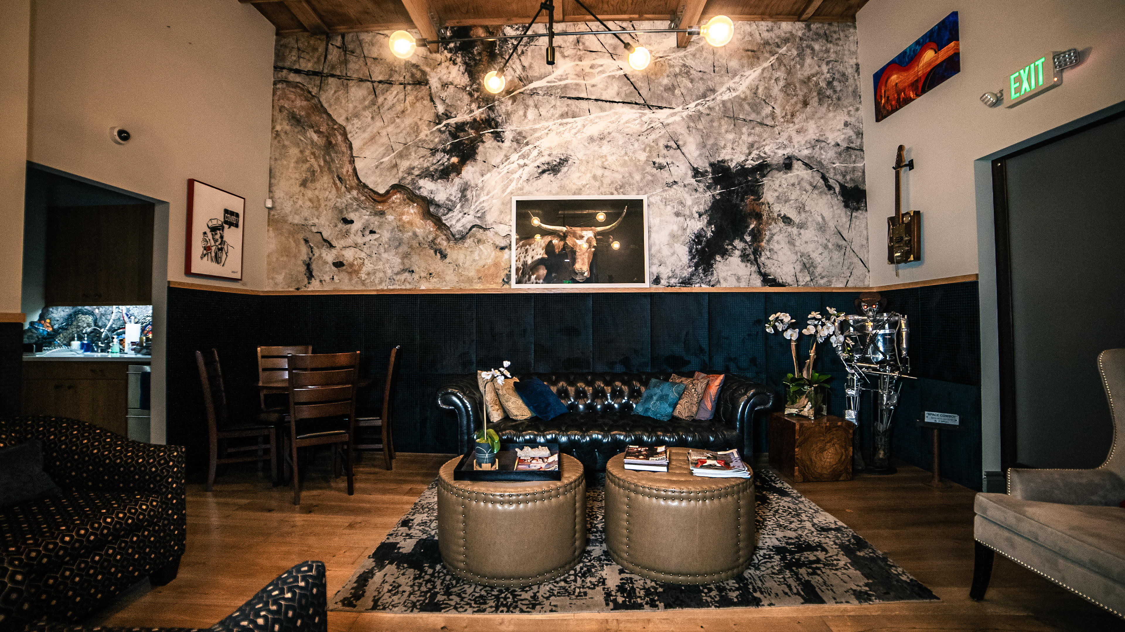 The image shows a stylish living area featuring a dark leather sofa, two round coffee tables, and a large stone wall with artwork, illuminated by pendant lights.