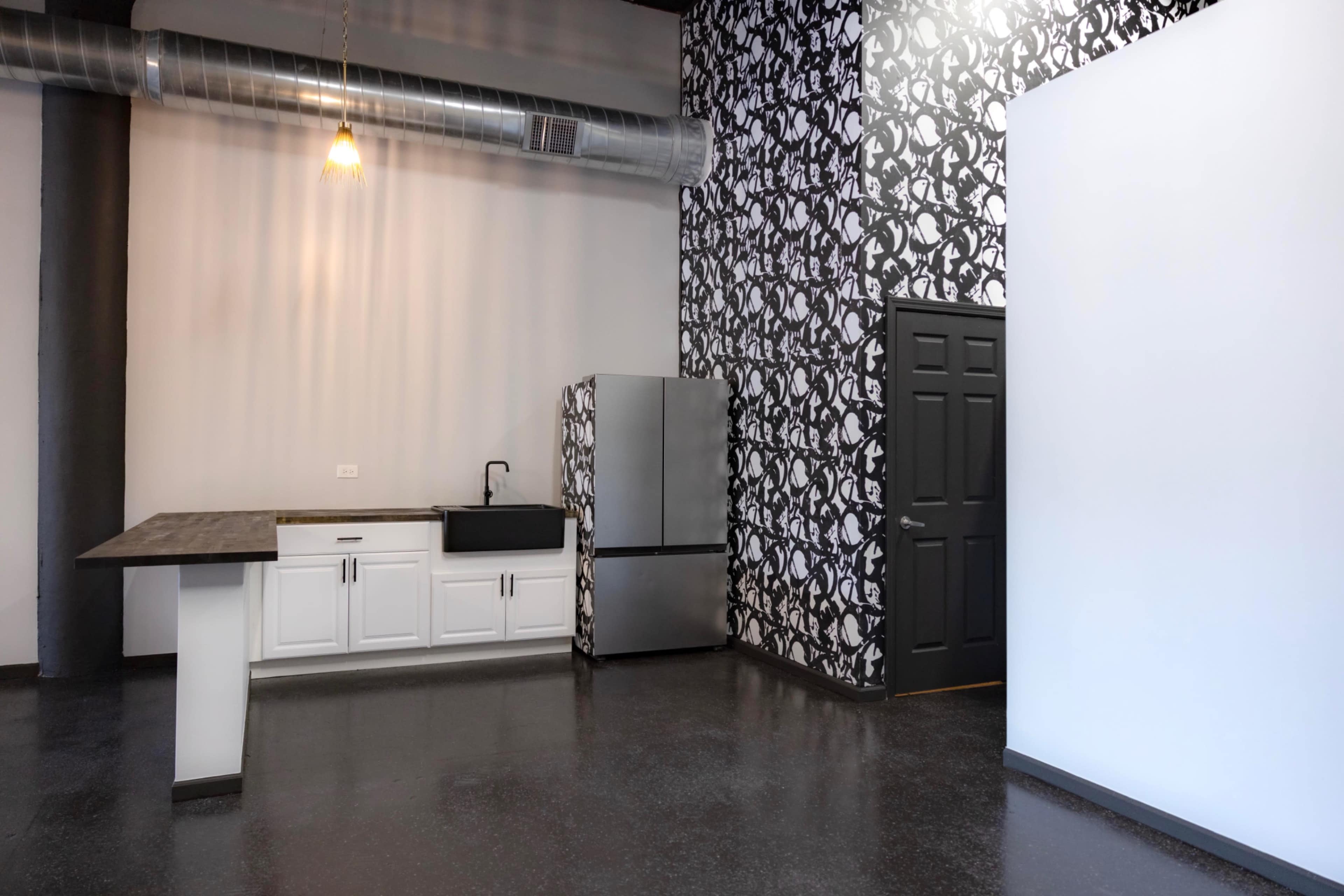 The image shows a modern kitchen area with a stainless steel refrigerator, a sink, and a counter, set against a patterned wallpaper and a black door leading to another space.