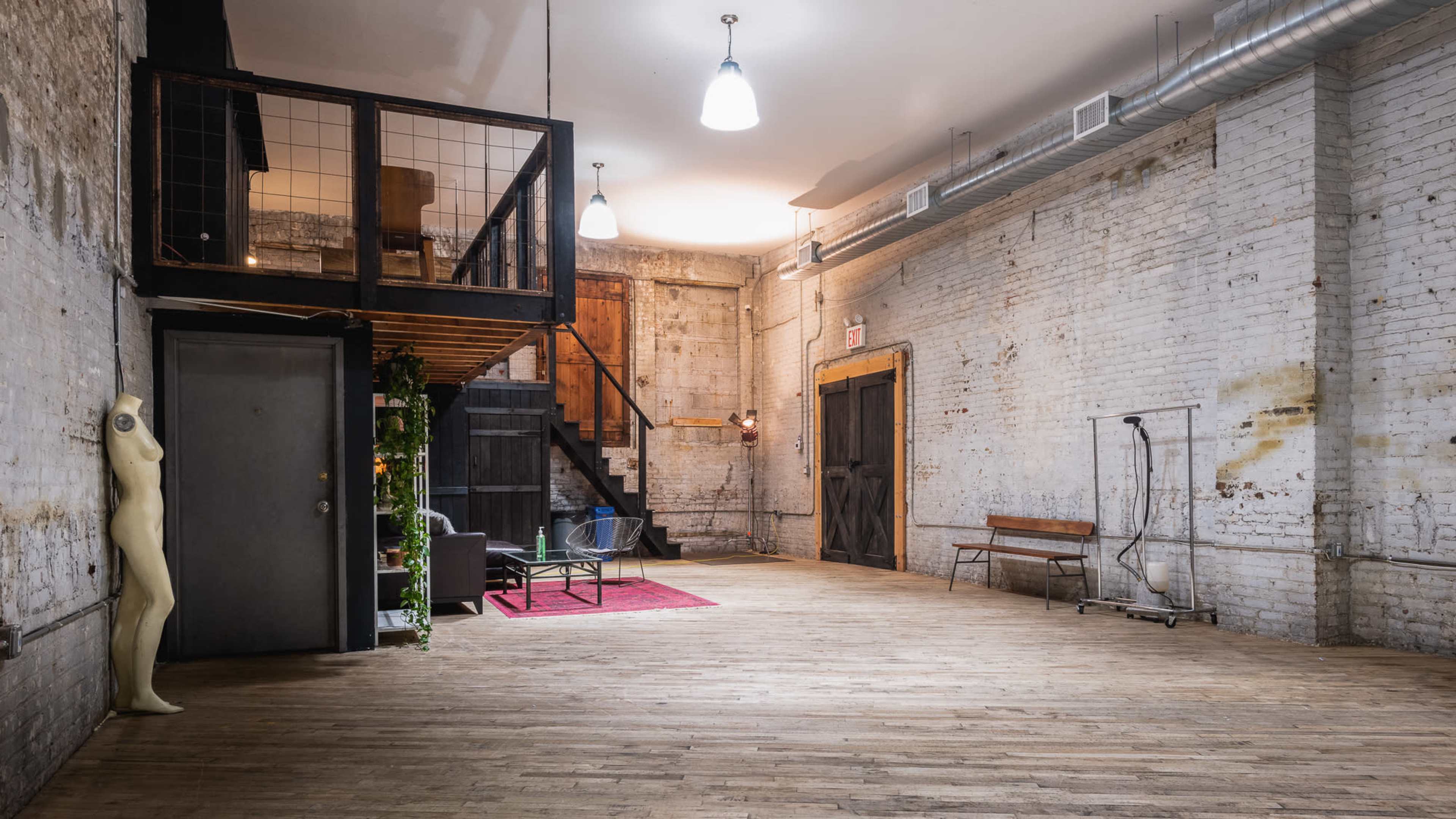 The image shows a spacious, industrial-style interior with exposed brick walls, wooden floors, a staircase, and minimal furniture.