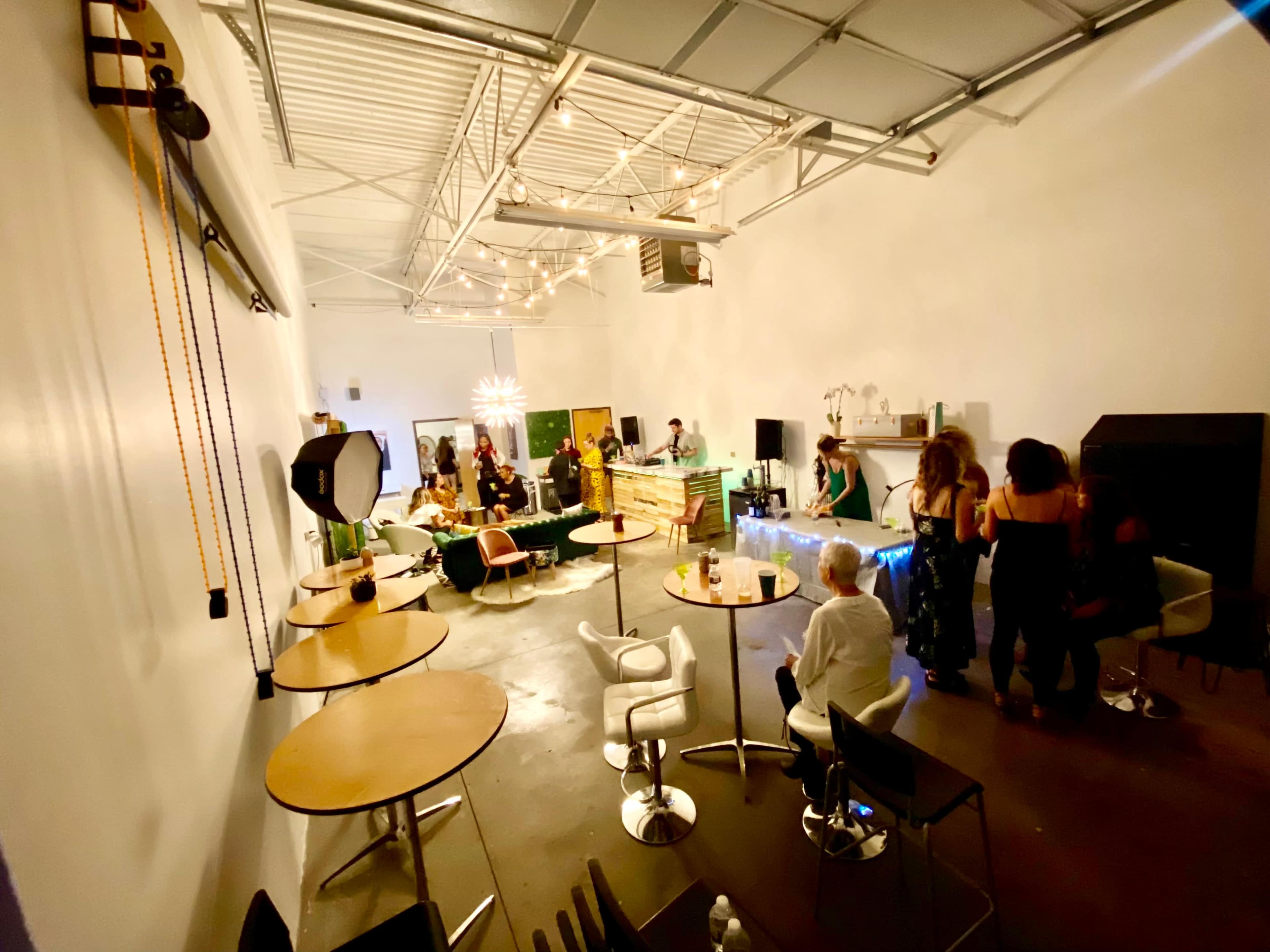 A spacious indoor venue features tables and seating, with people socializing and a bar area in the background.