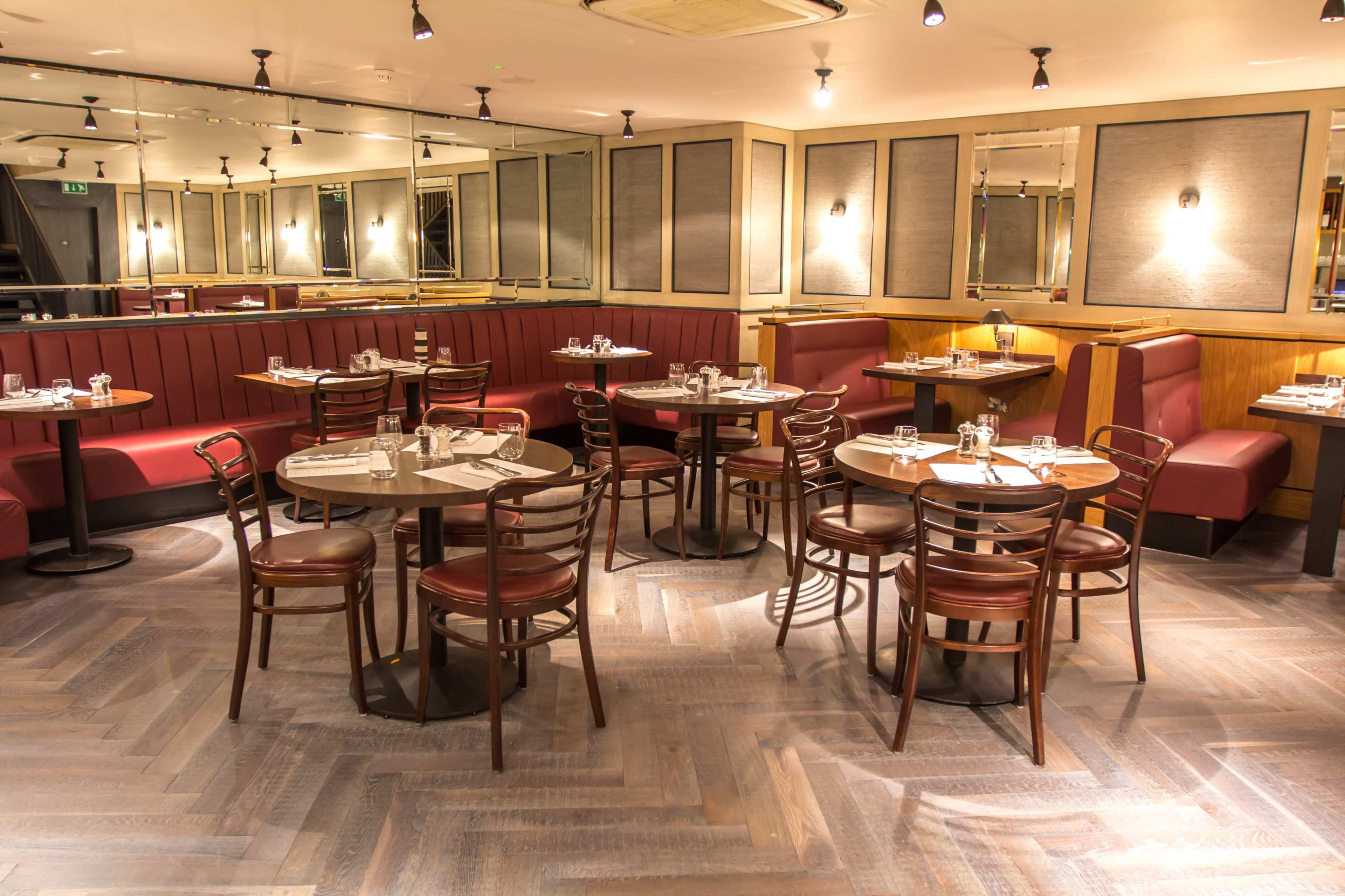 The image shows a well-lit restaurant interior with several round tables, chairs, and red upholstered booths arranged on a wooden floor.