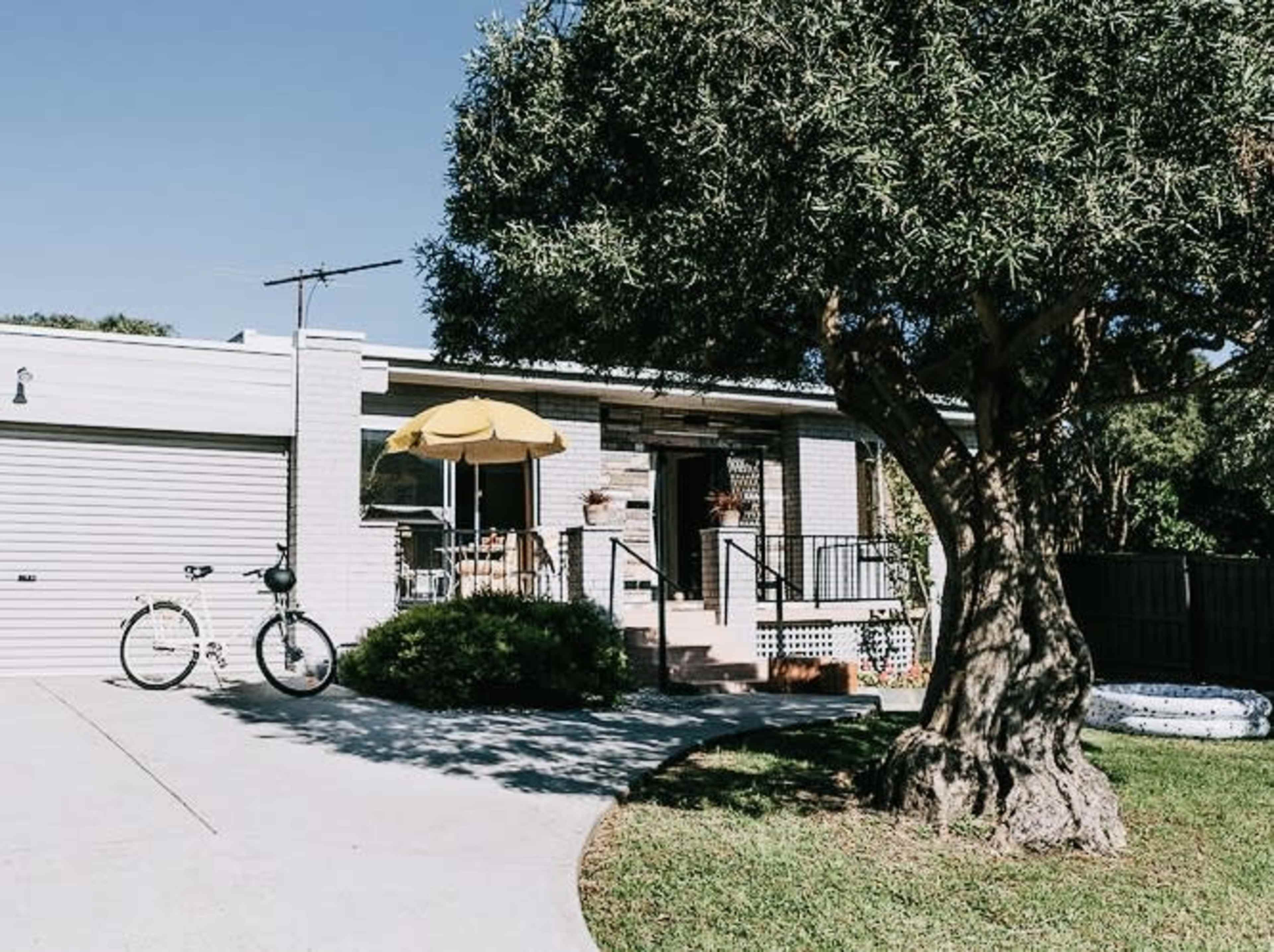 A small, white house with a yellow umbrella over a seating area in the front yard, flanked by a bicycle and greenery.
