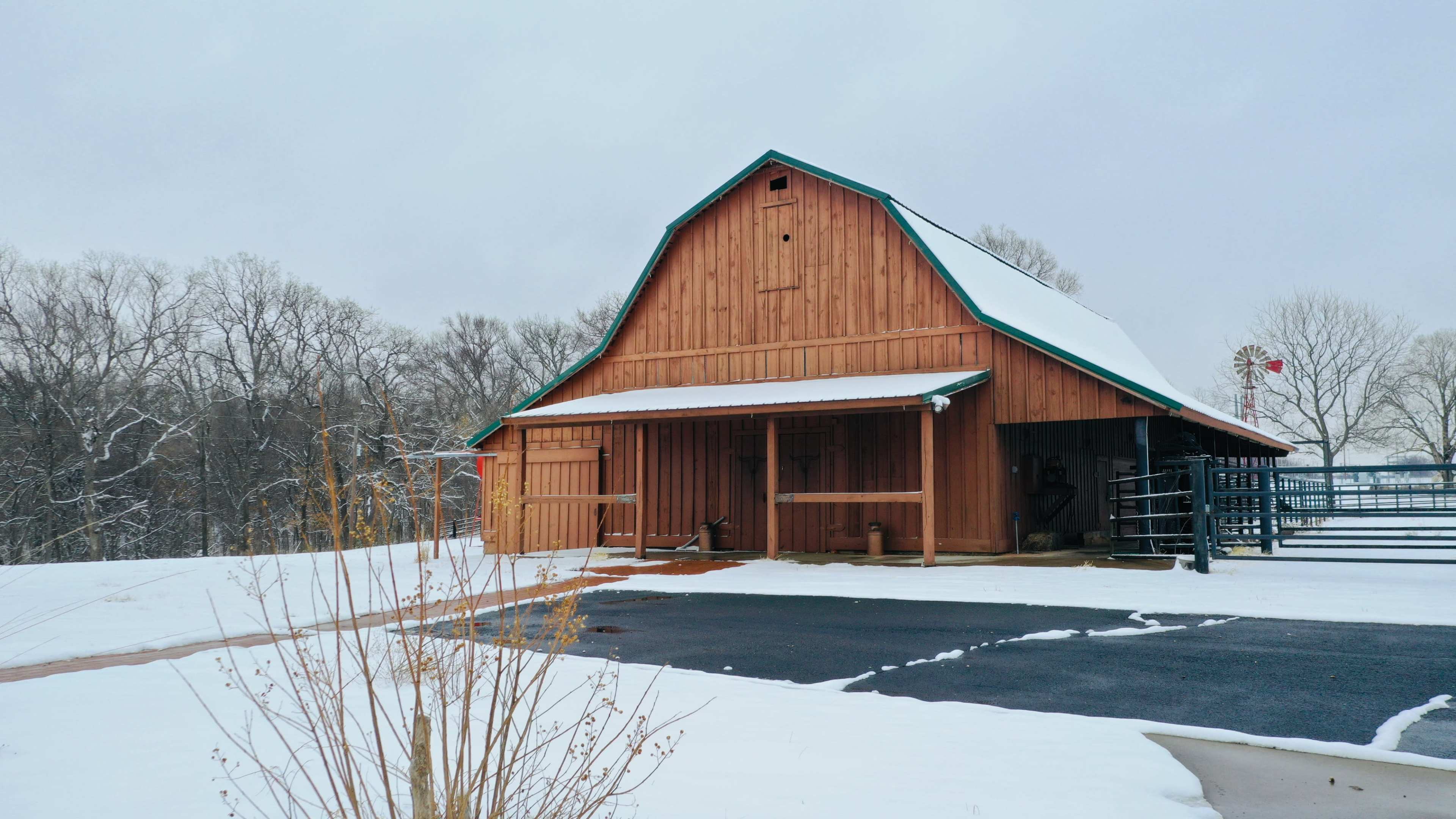 A wooden barn with a green roof stands beside a snow-covered driveway in a rural area.