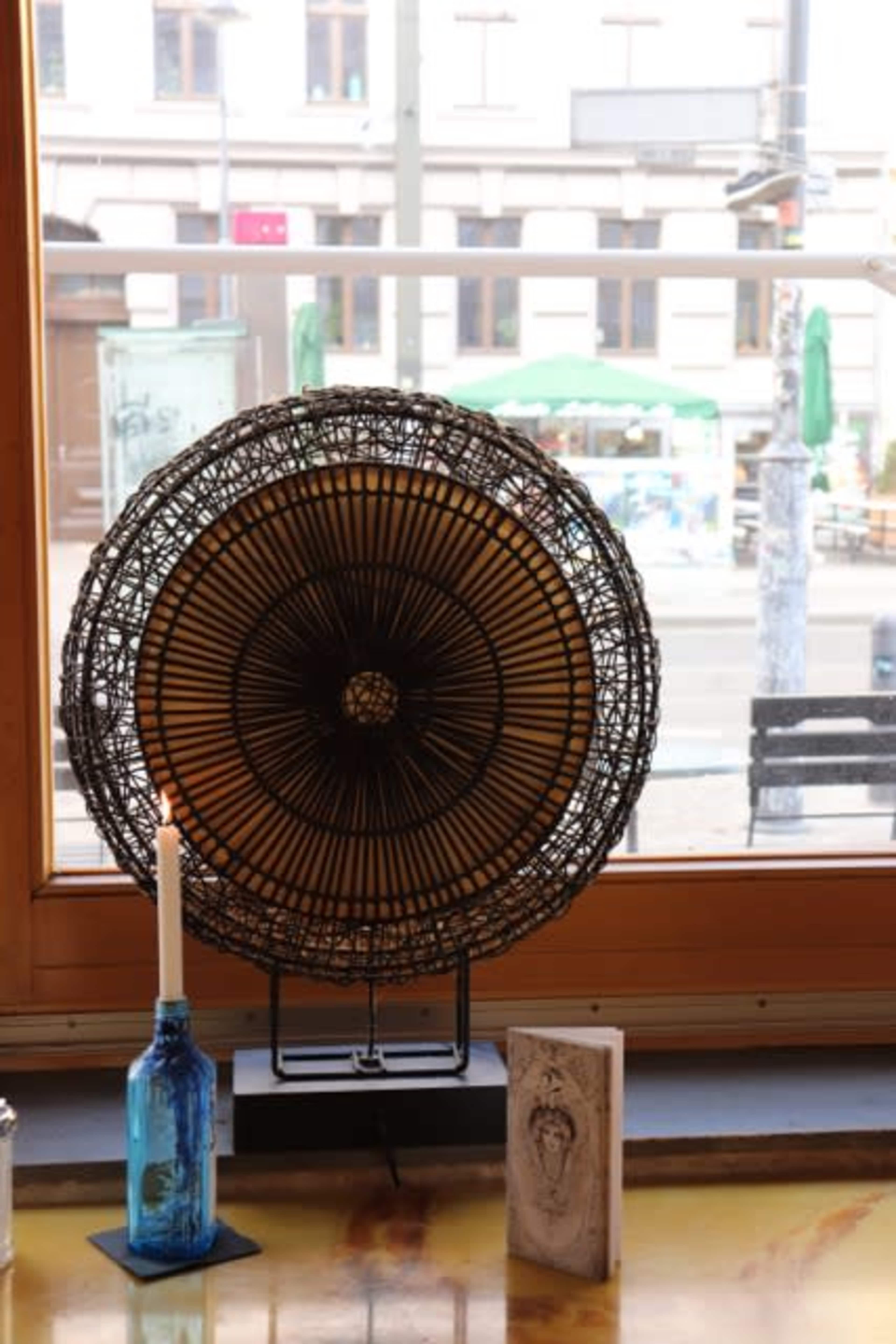 A decorative fan and a blue glass bottle sit on a wooden table in front of a large window with a view of street activity outside.