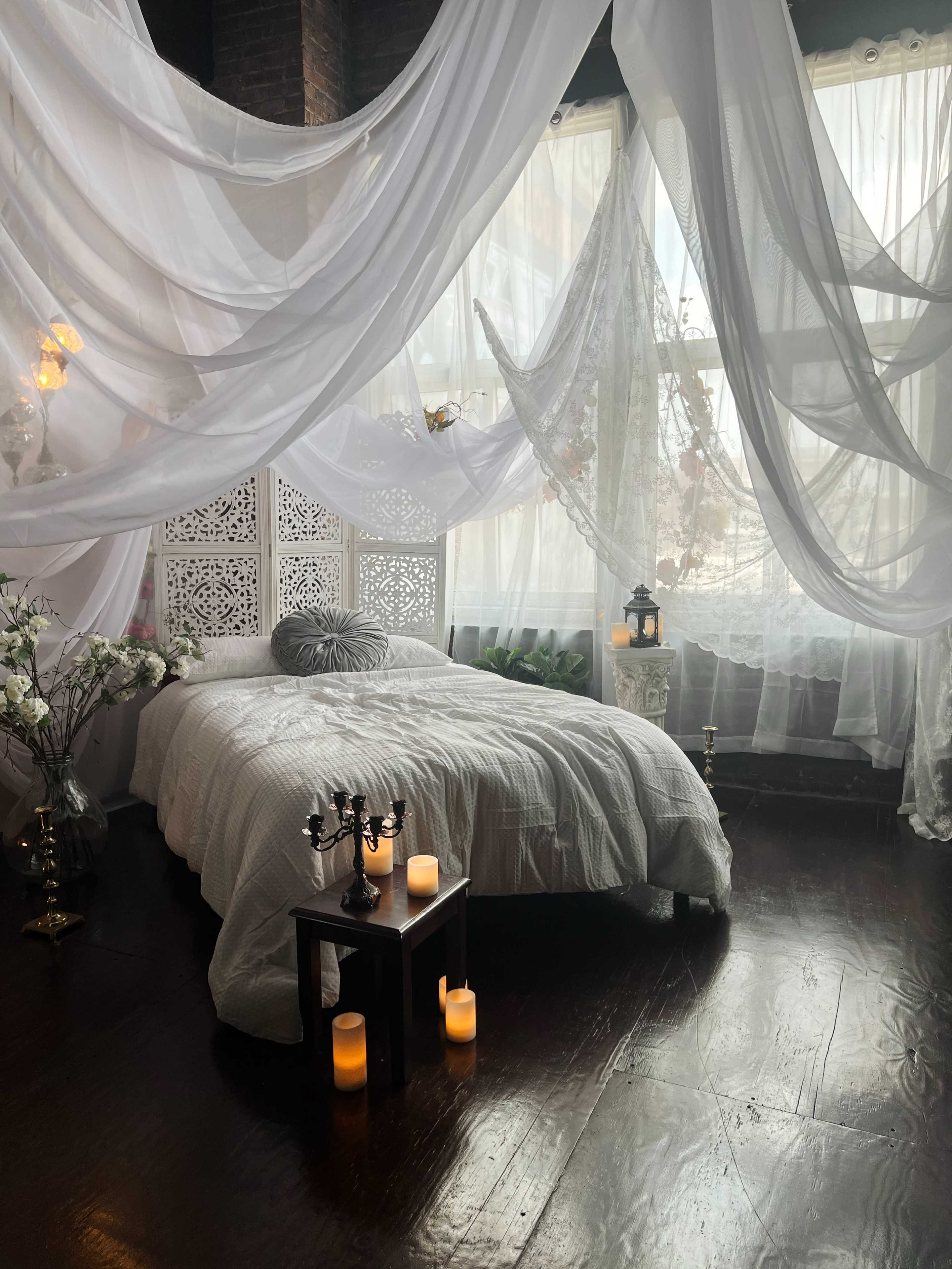 A bed with a light-colored quilt is surrounded by flowing sheer fabric and candles, set in a softly lit room with decorative plants and intricate wooden screens.