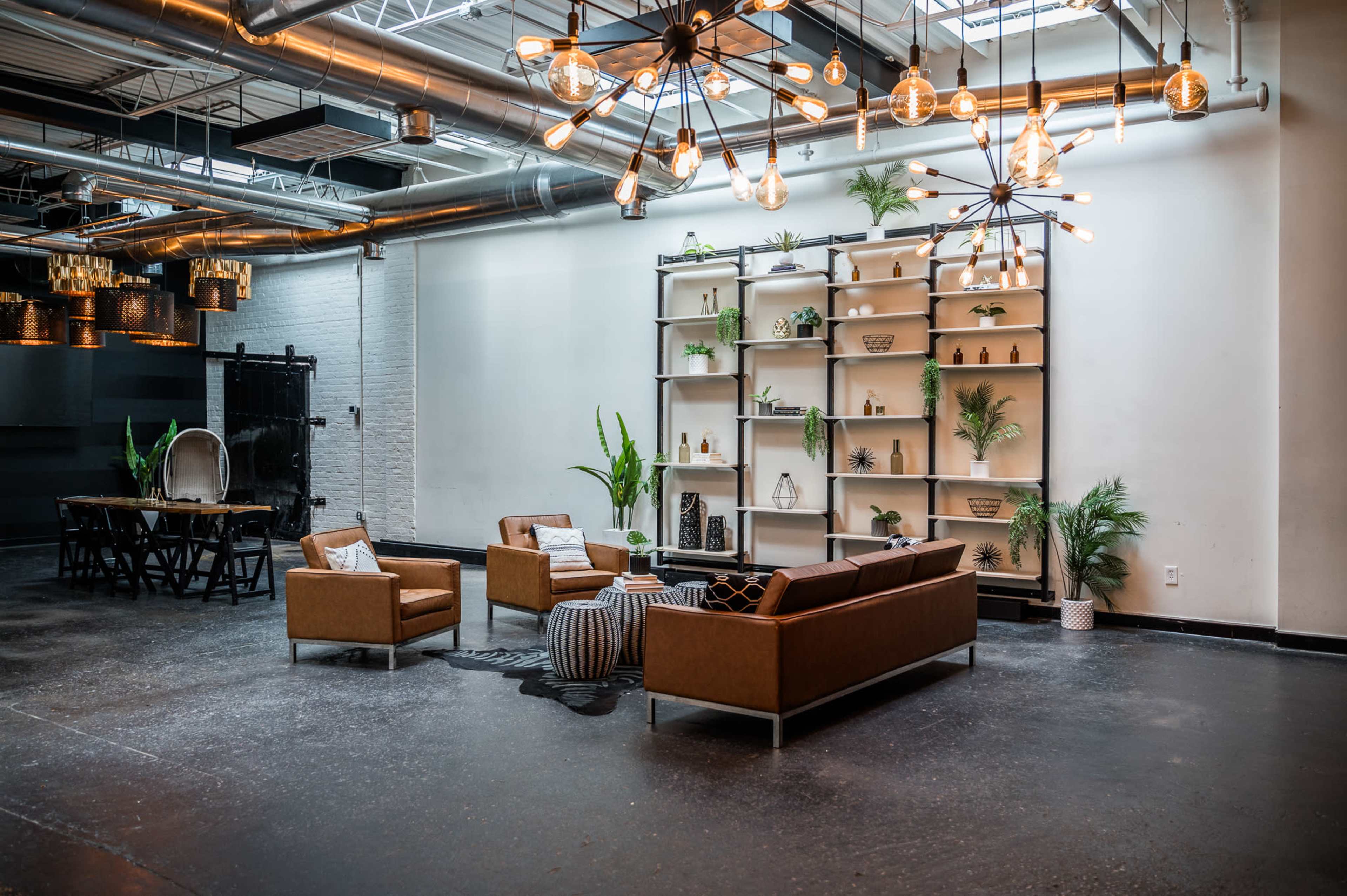 A modern industrial-style interior with a seating area featuring a brown leather couch, two armchairs, and a coffee table, alongside a decorative shelf and a dining table in the background.
