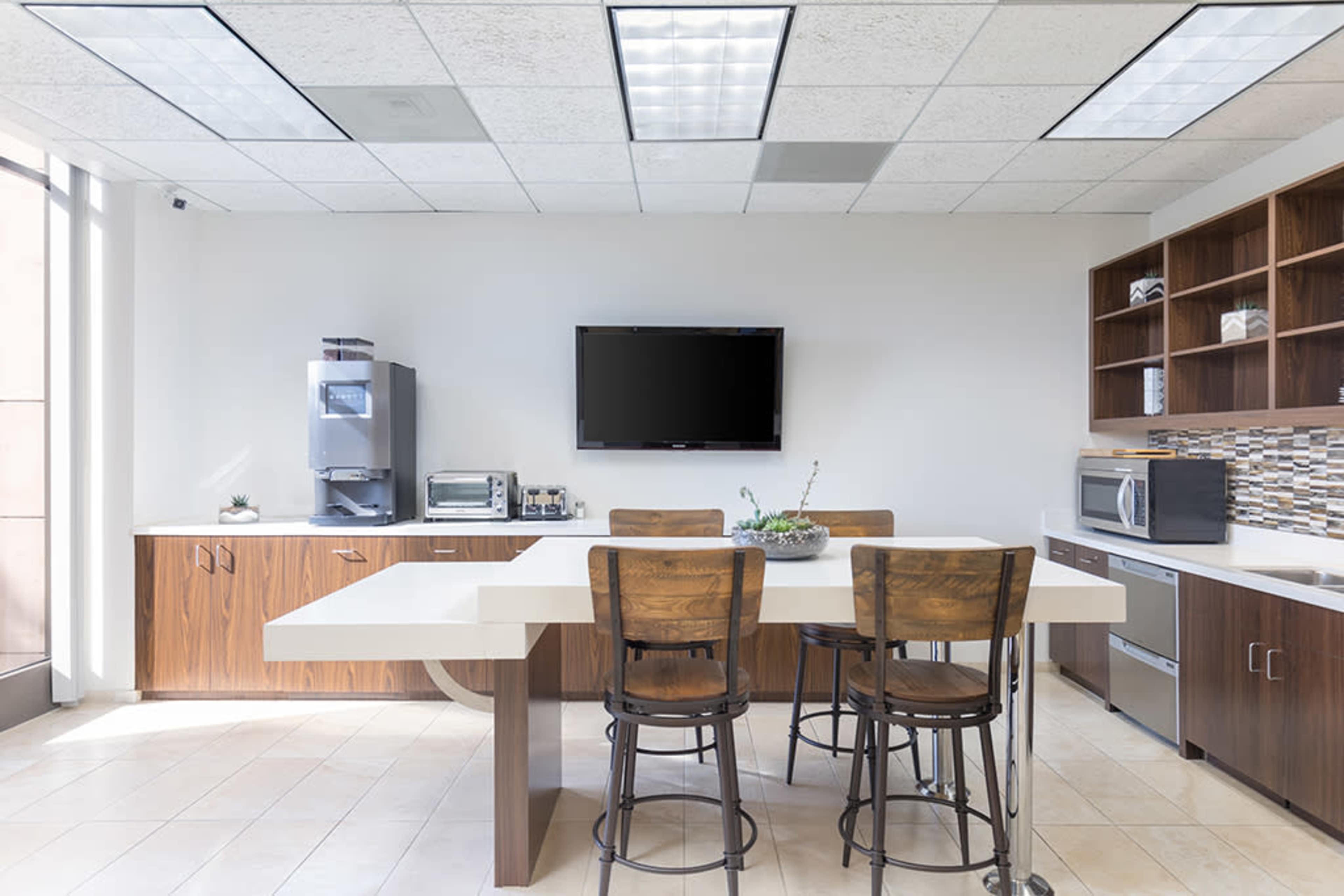 The image shows a modern kitchen area with a large white table, wooden stools, and various appliances, including a microwave and a water dispenser, against a light-colored wall.