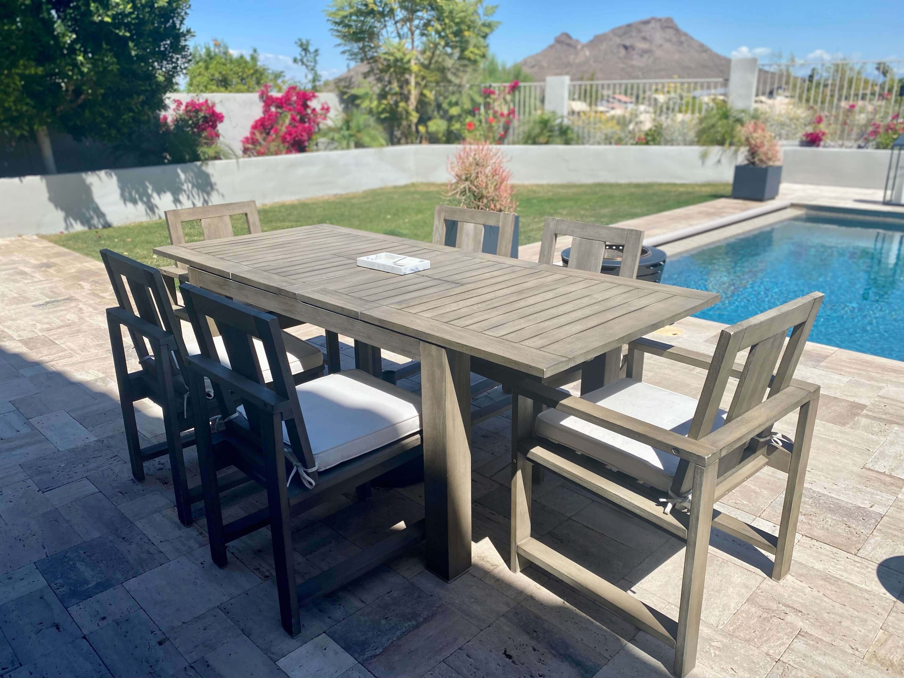 A wooden dining table with eight chairs is set beside a swimming pool, surrounded by greenery and colorful flowers in a residential backyard.
