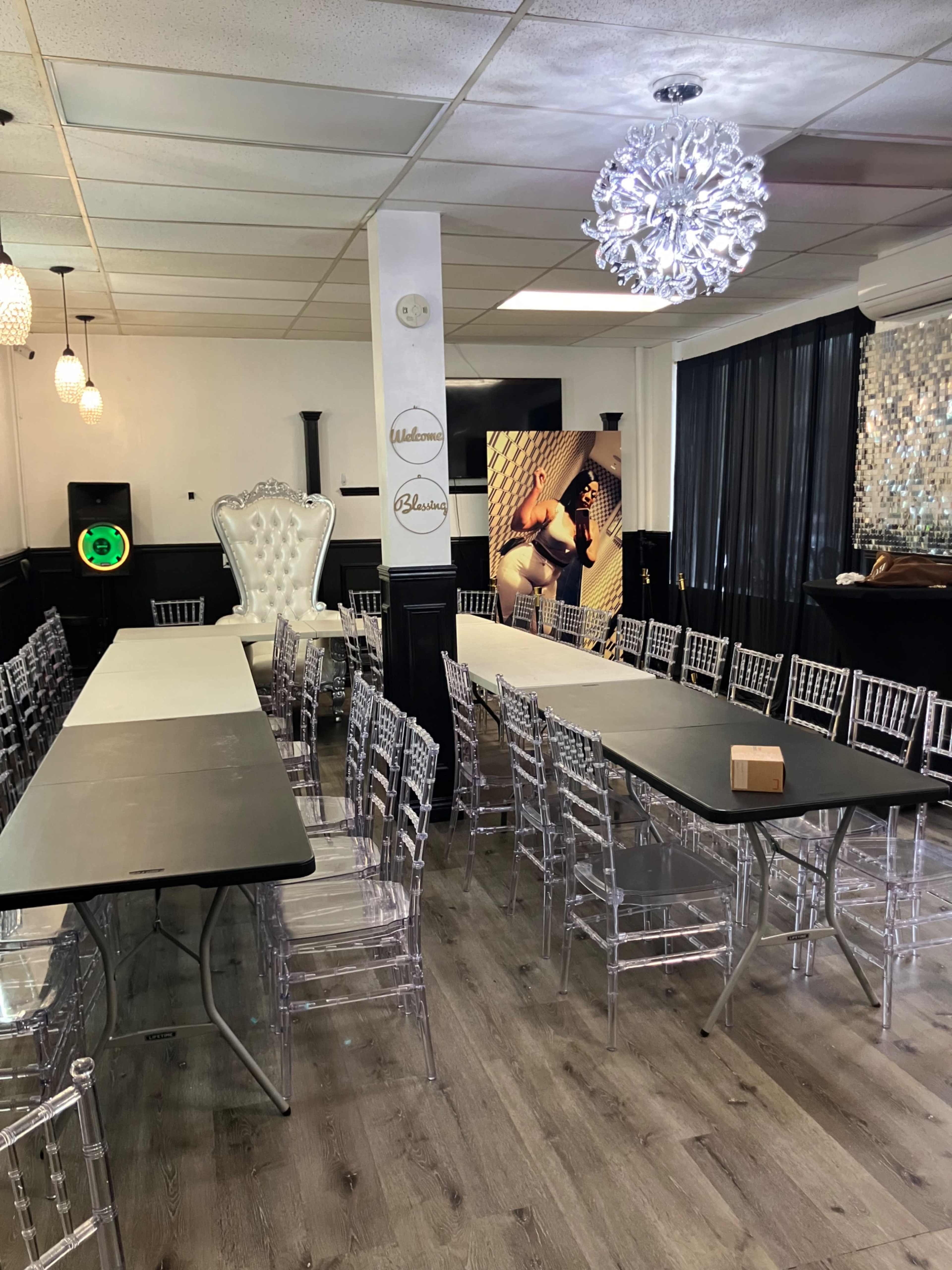 The image shows a spacious event room set up with multiple rows of tables and clear acrylic chairs, featuring decorative lighting and large windows.