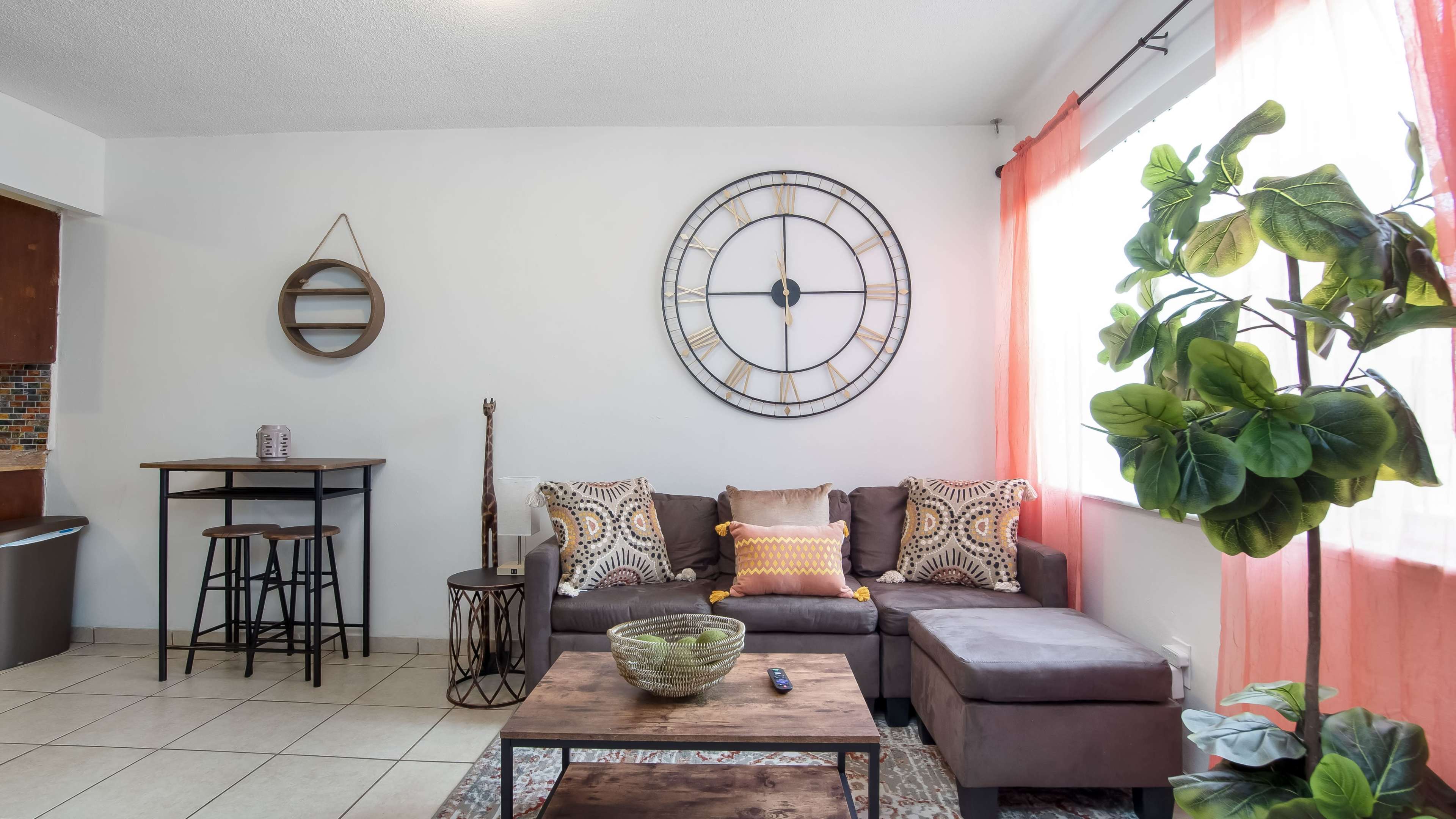 A living room with a brown couch, decorative pillows, a large wall clock, and plants, accompanied by a small dining table and chairs.