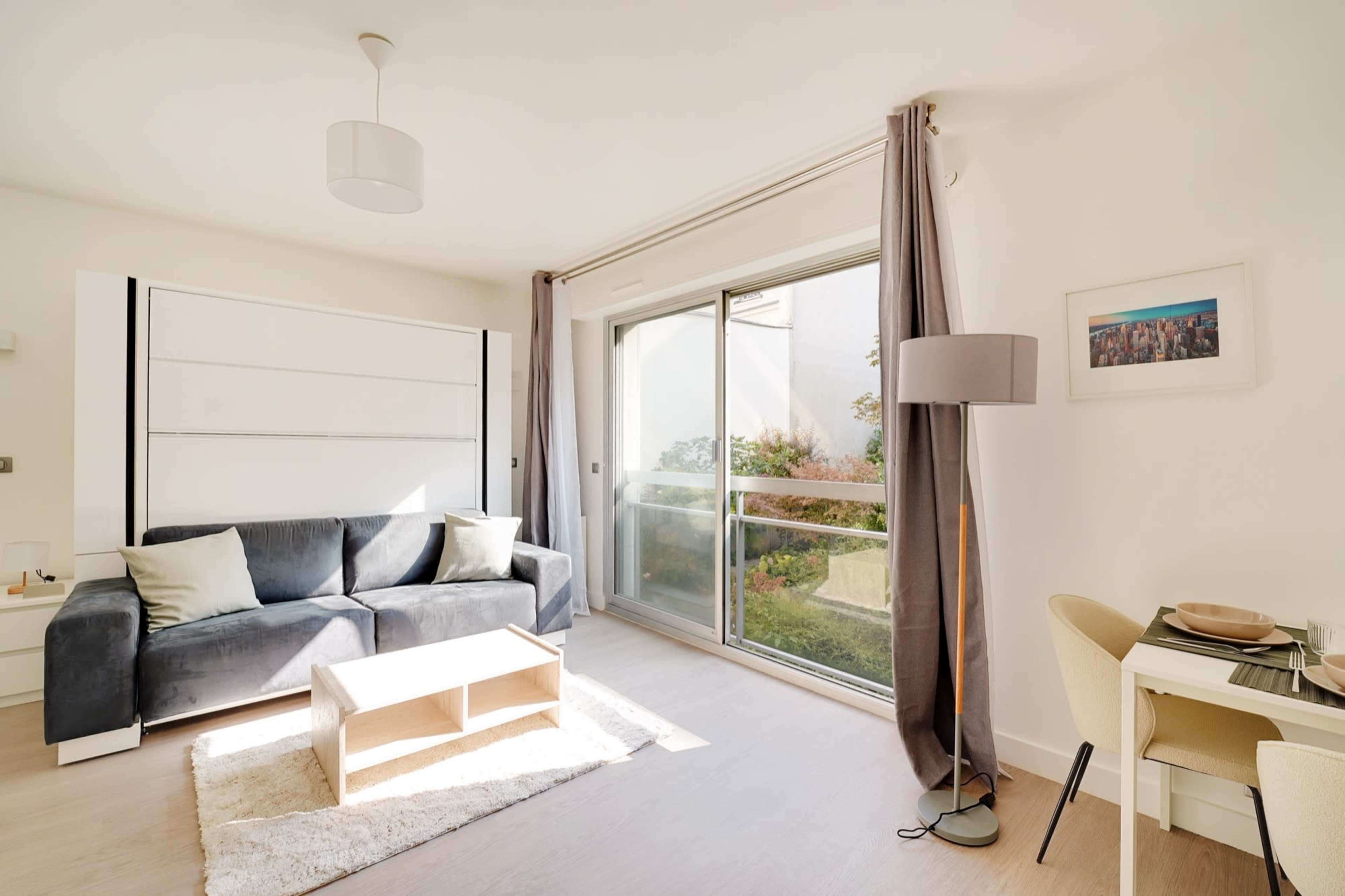 The image shows a modern living room with a gray sofa, a small wooden table, a floor lamp, and a glass door leading to an outdoor area.