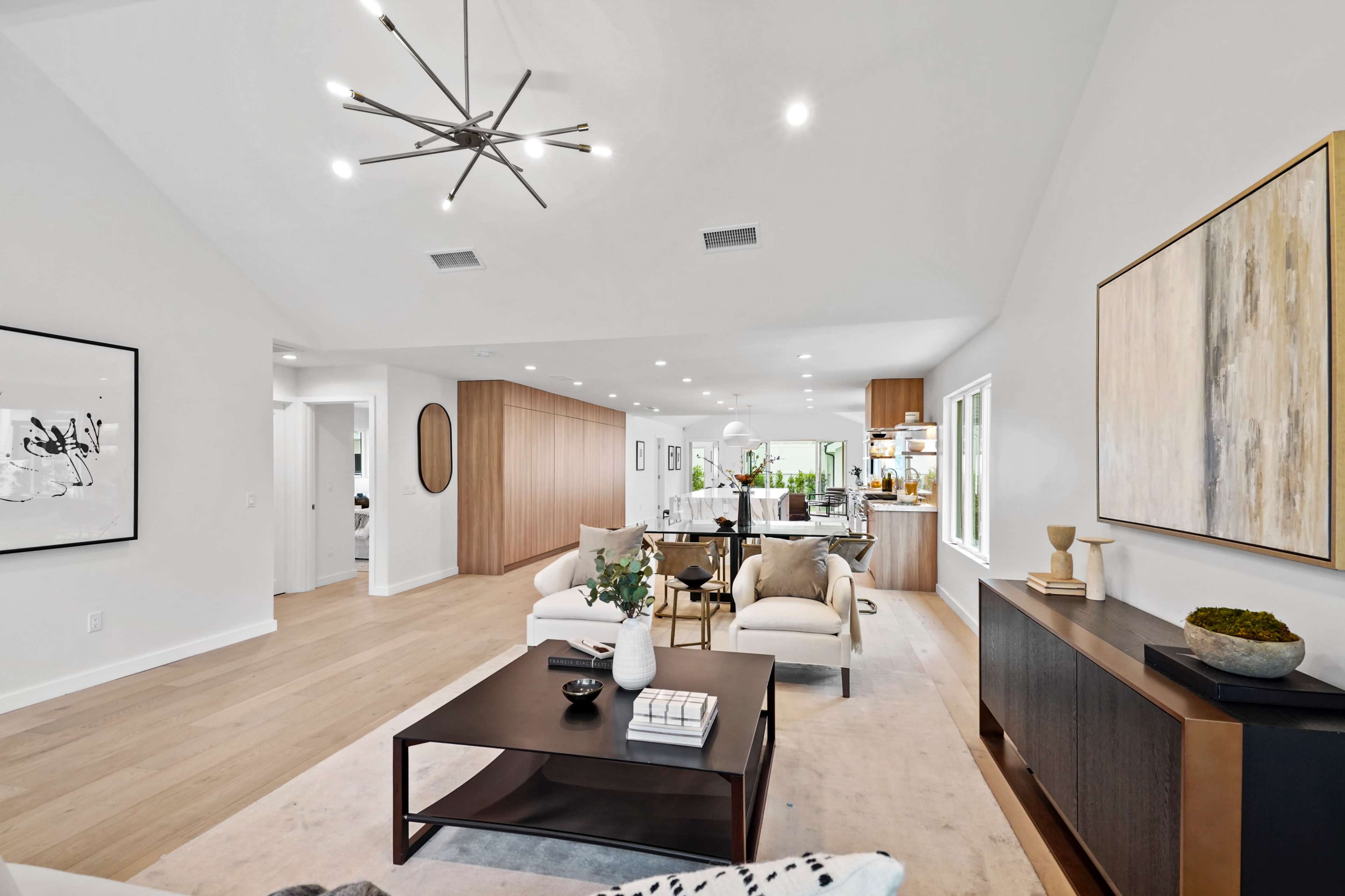 A modern open-concept living area with light-colored walls, hardwood flooring, and a kitchen visible in the background.