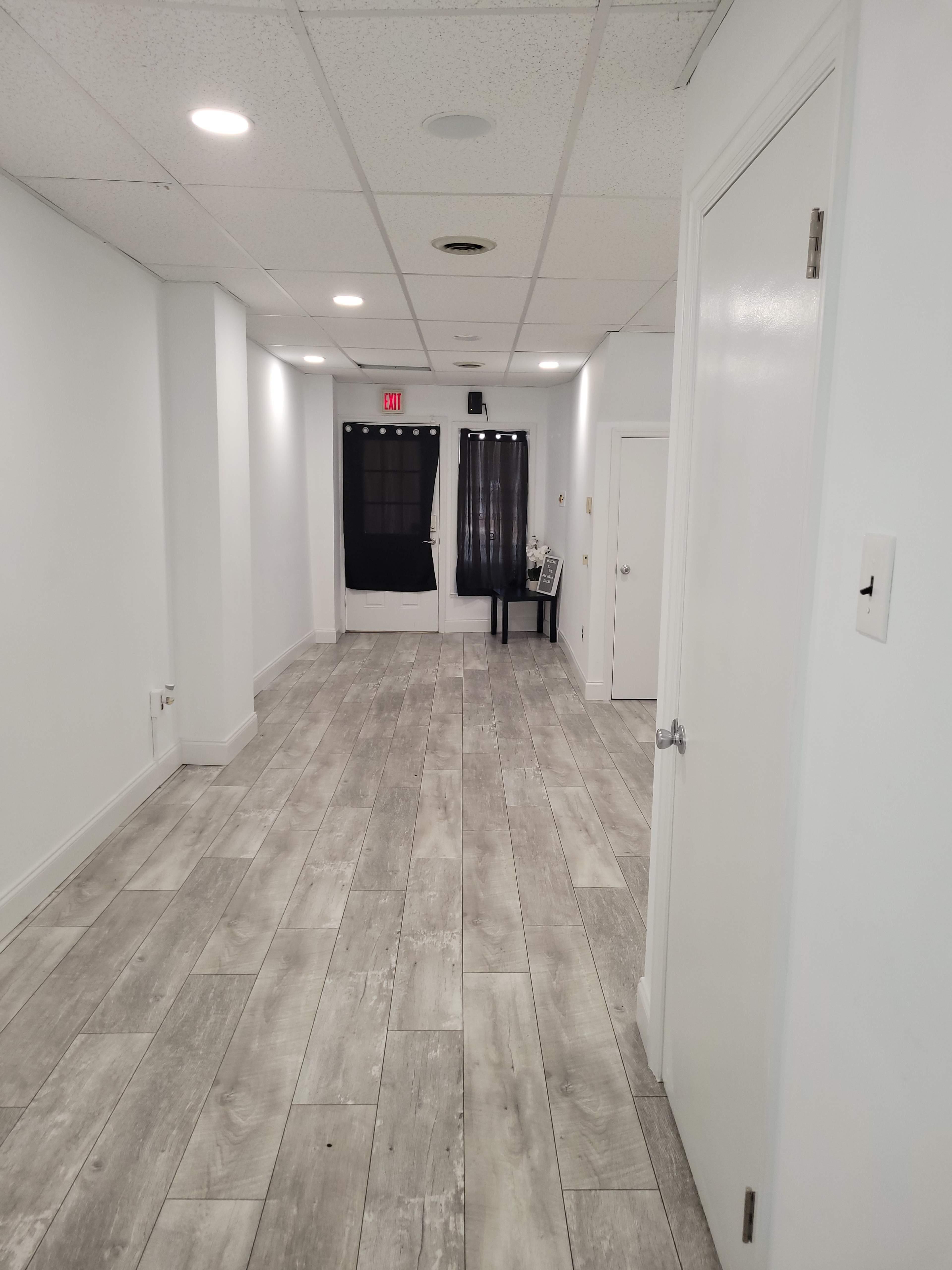 The image shows a long, empty hallway with gray tiled flooring, white walls, and a closed door at one end.