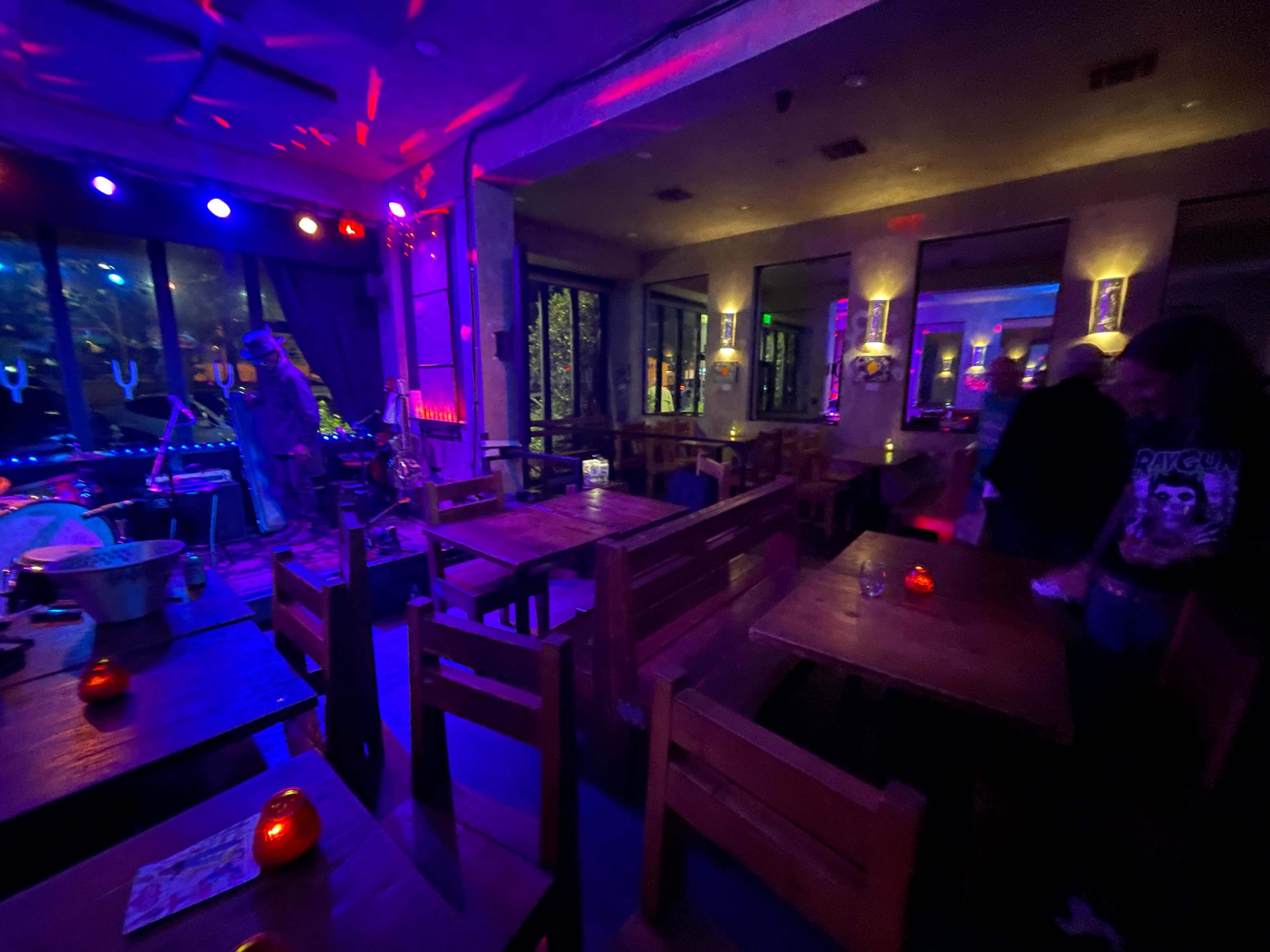 The image shows a dimly lit bar or restaurant with wooden tables, colorful lights, and a small stage set up for a performance.